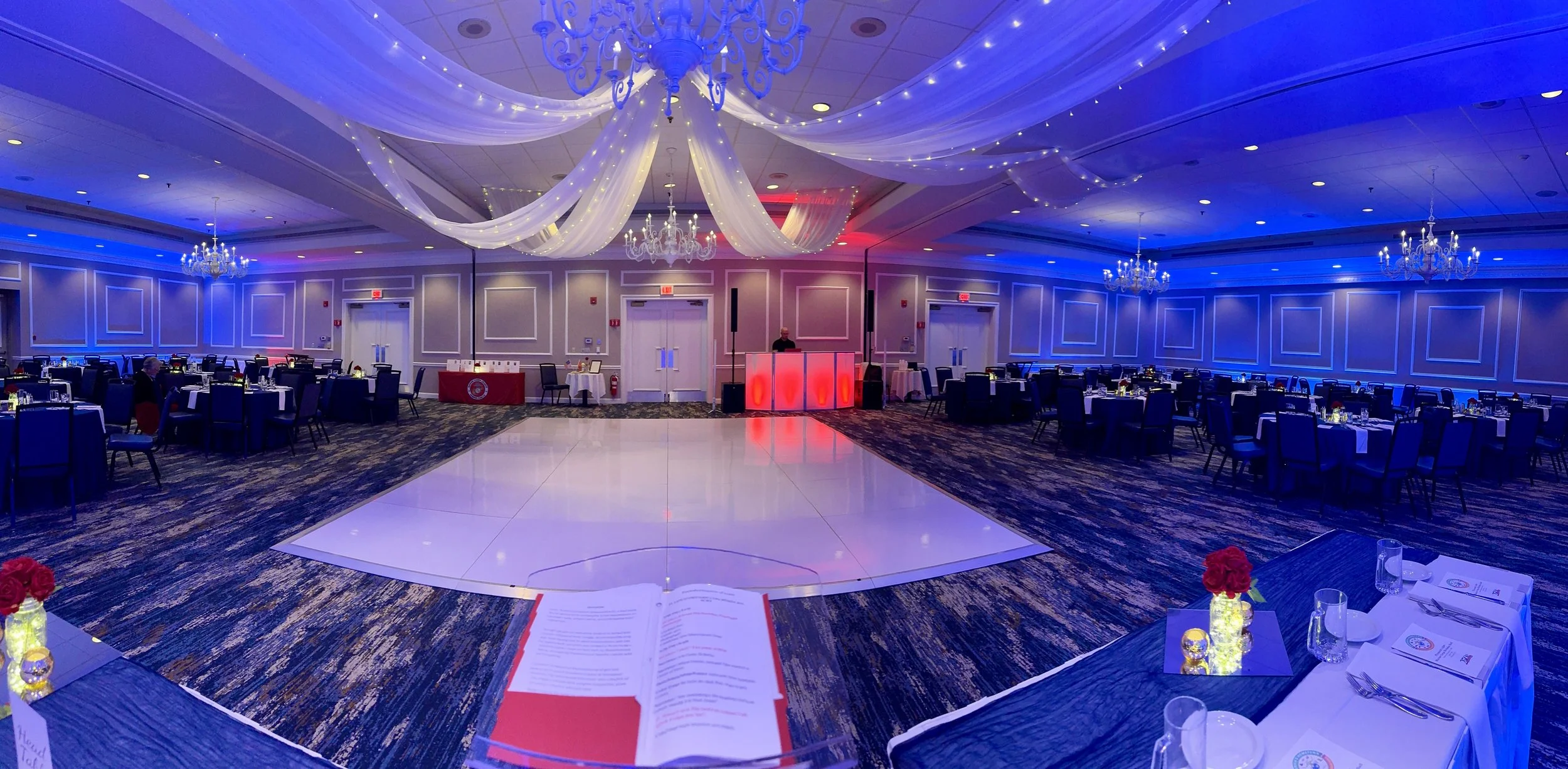 An elegant banquet hall decorated for a wedding reception with a central dance floor, white tent-like drapes with string lights, chandeliers, round tables with black chairs, and table settings with flower centerpieces.