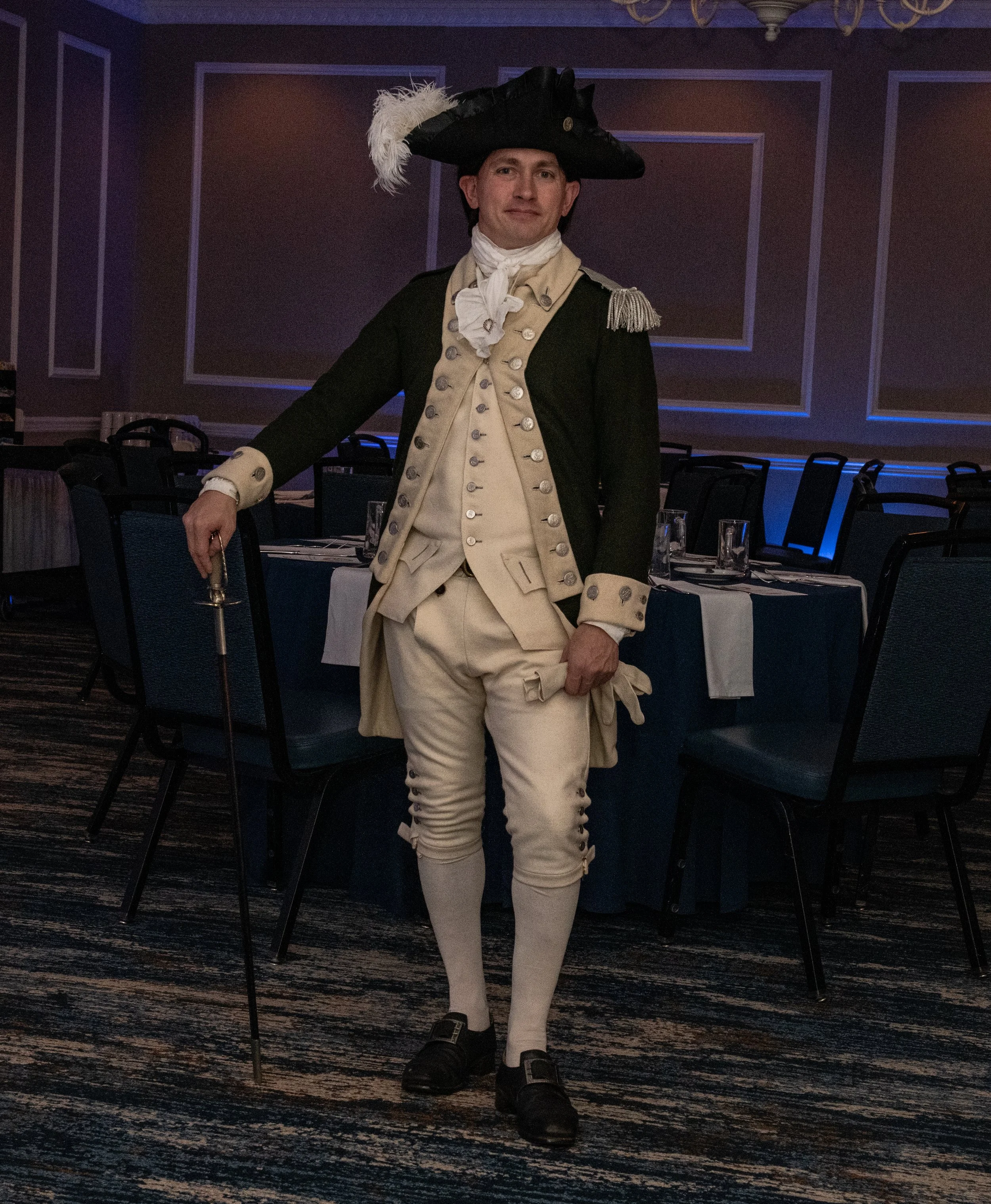A man dressed as a historical figure, possibly from the 18th century, standing in a dimly lit banquet hall. He is wearing a black tricorn hat with a white feather, a beige waistcoat with metal buttons, a matching beige breeches, a long dark coat with