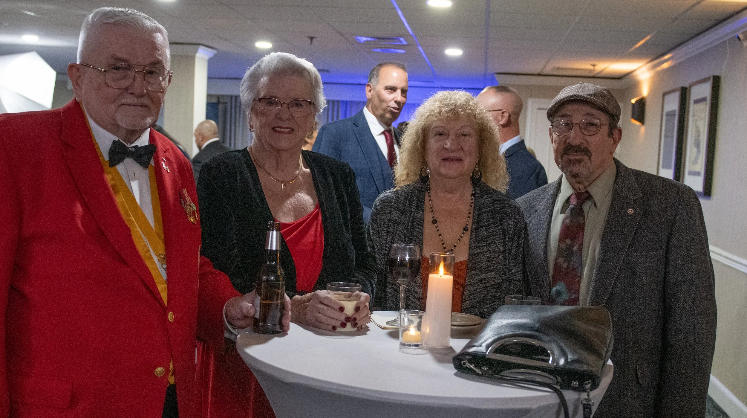 Group of five older adults at a social gathering, standing around a tall table with drinks and a candle, in an indoor event space with framed pictures on the wall.