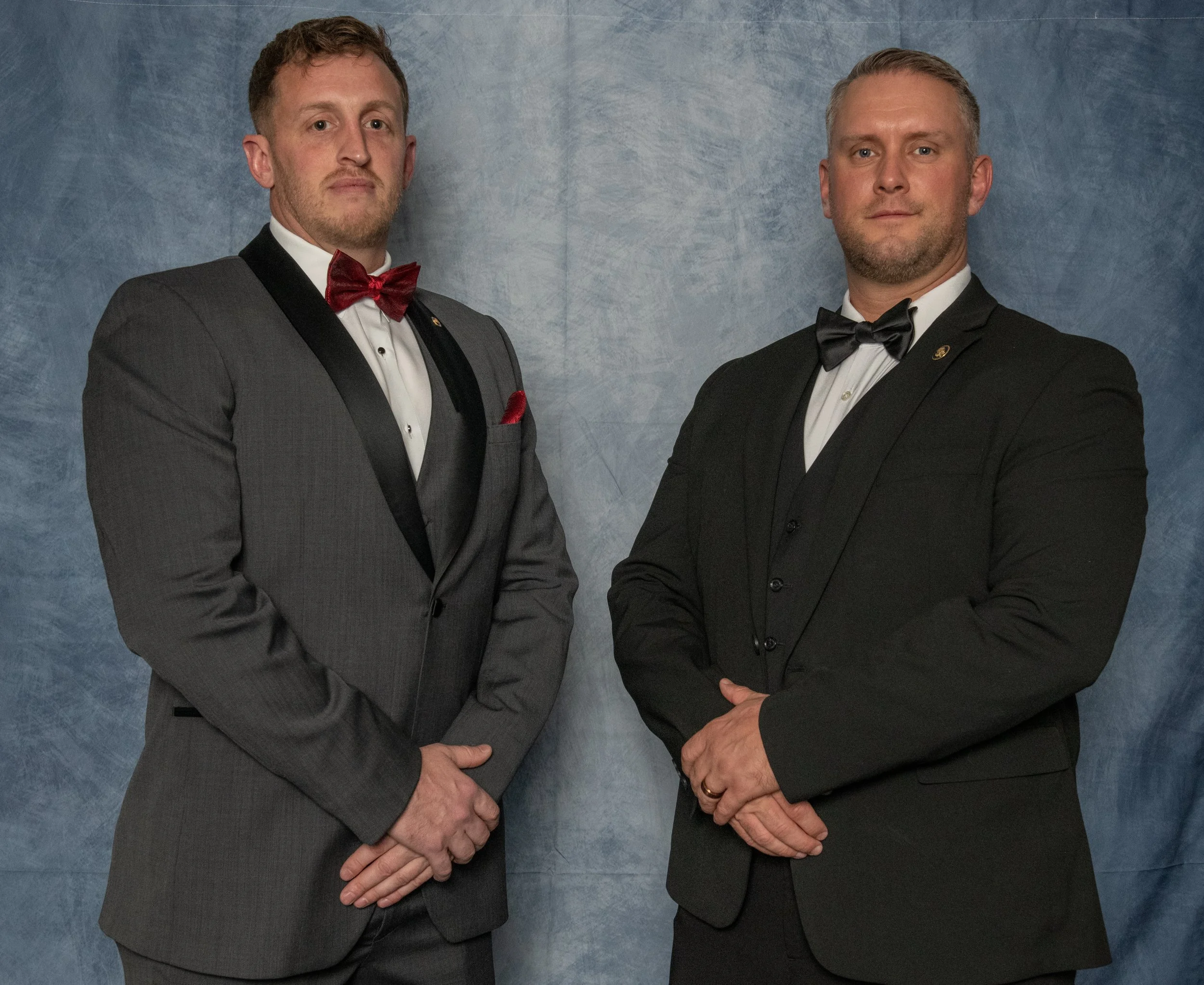 Two men in formal tuxedos with bow ties and cummberbunds standing with hands clasped in front of a blue-gray backdrop.