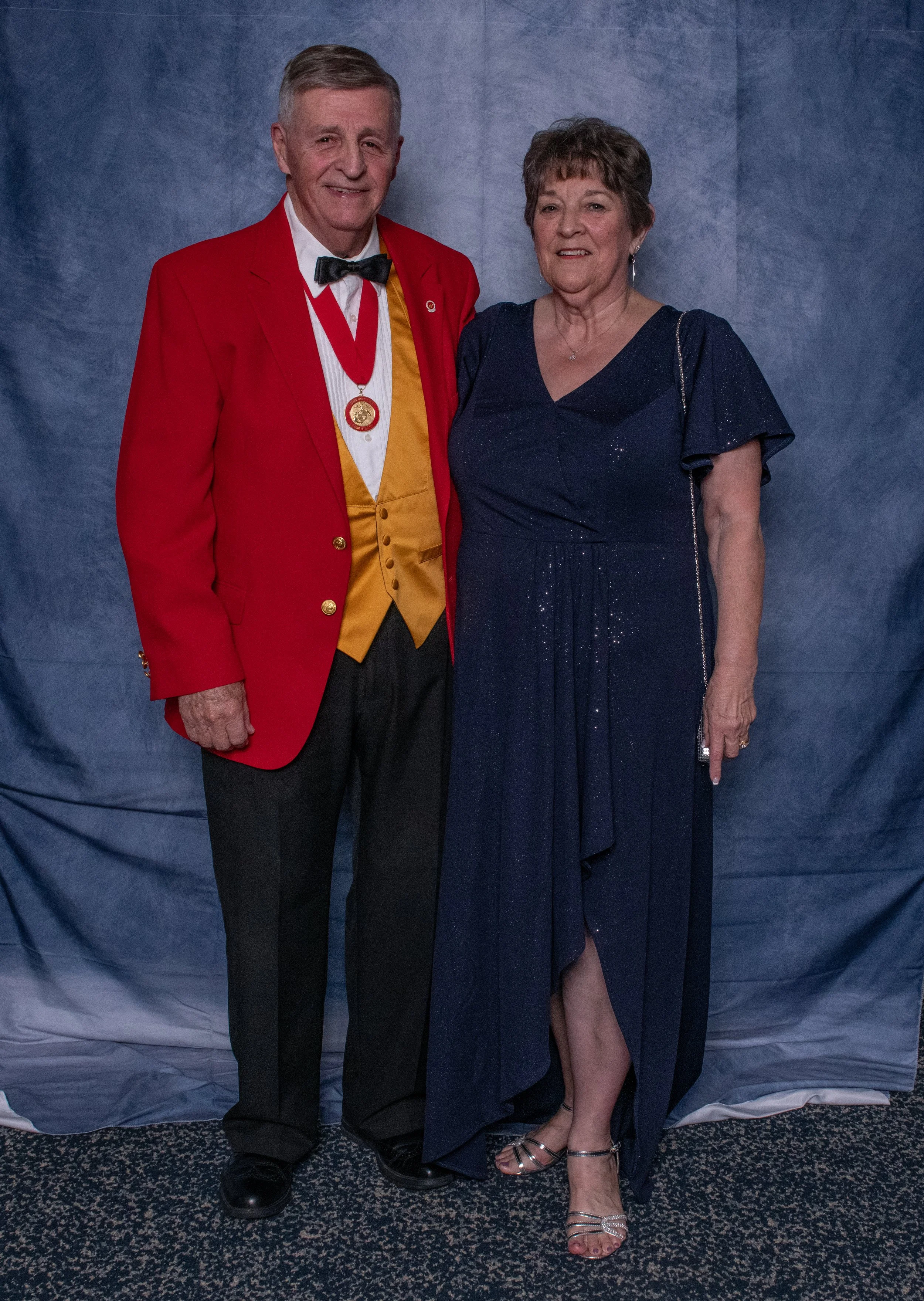 An elderly man in a red tuxedo jacket, yellow vest, white shirt, black bowtie, and a medal, standing next to an elderly woman in a navy blue evening gown with glitter accents, both smiling in front of a blue backdrop.