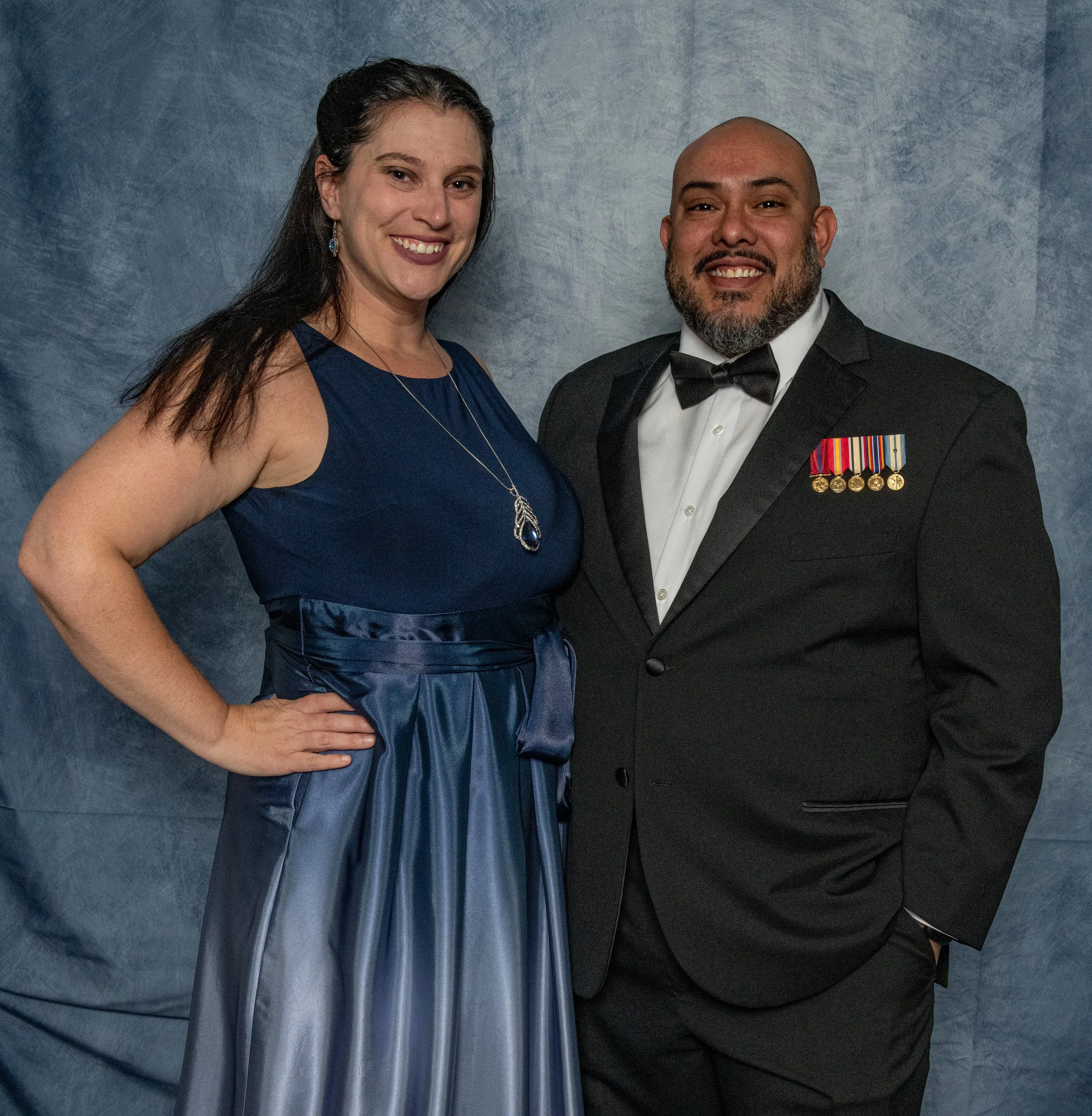 A woman in a blue satin evening gown and a man in a black tuxedo with military medals, both smiling and posing together against a gray backdrop.