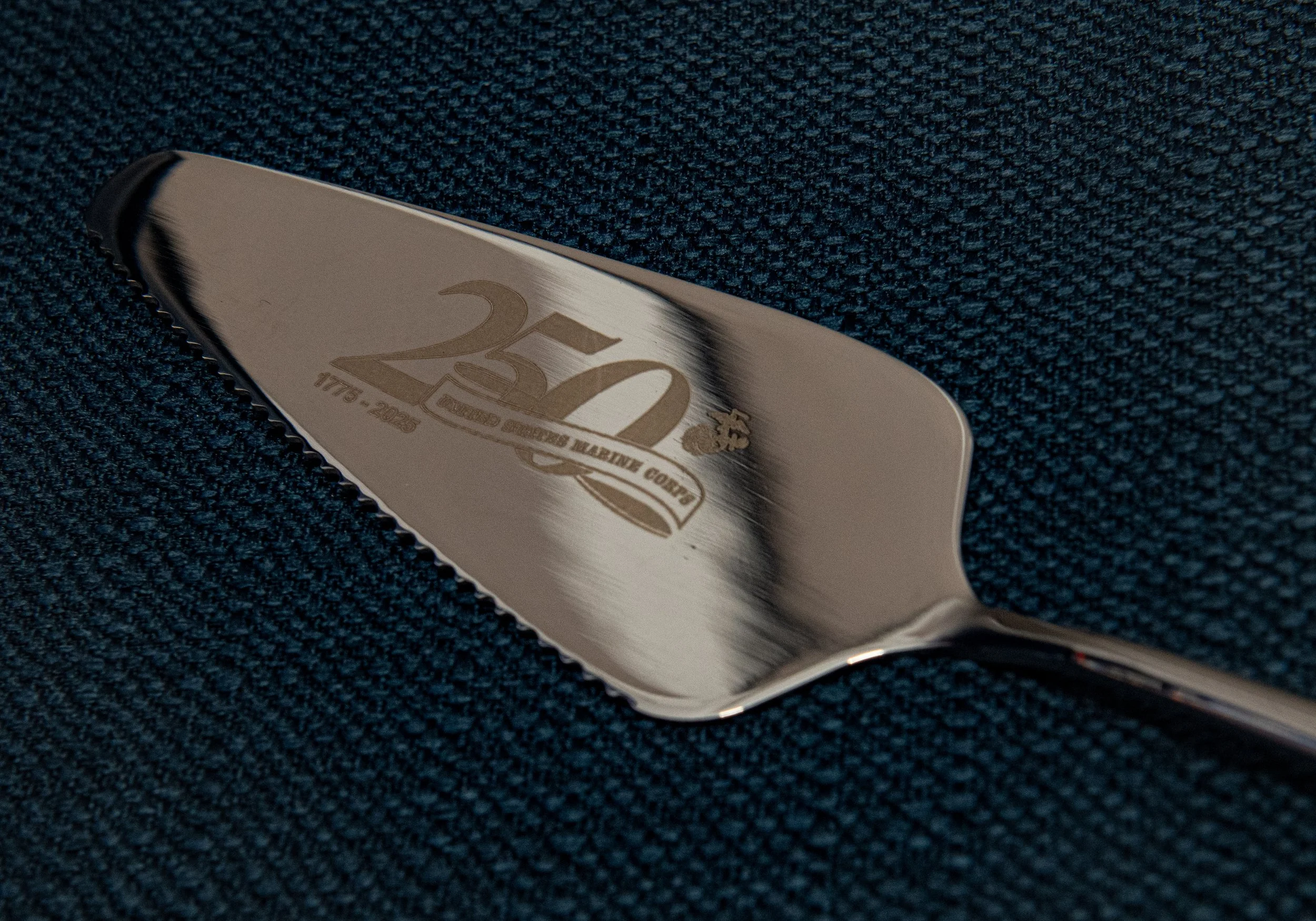 A metal spatula with engraving celebrating 250 years of the United States Marine Corps, lying on dark textured fabric.
