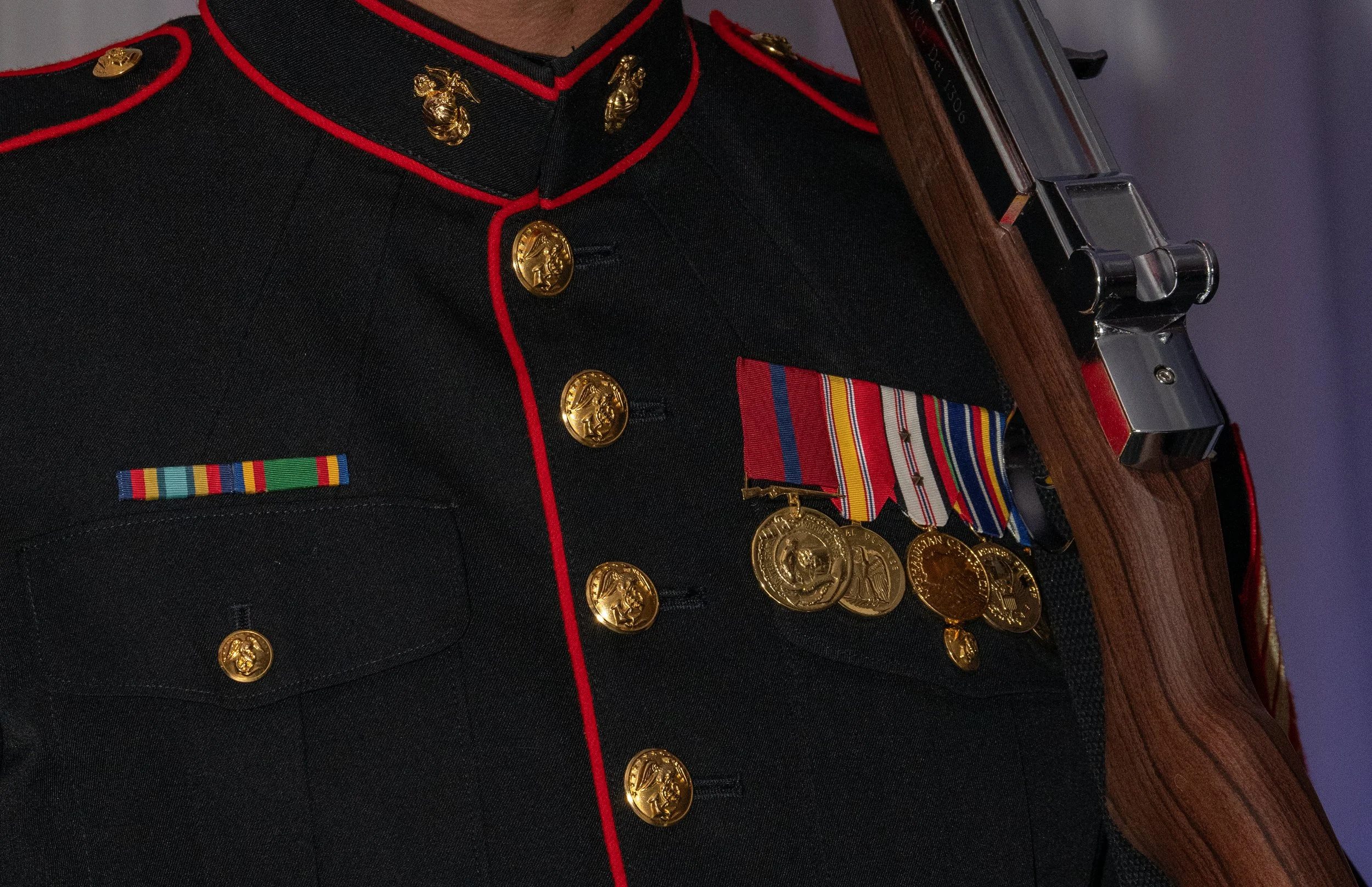 A person in a military dress uniform with gold buttons, red piping, and medallions, holding a rifle.