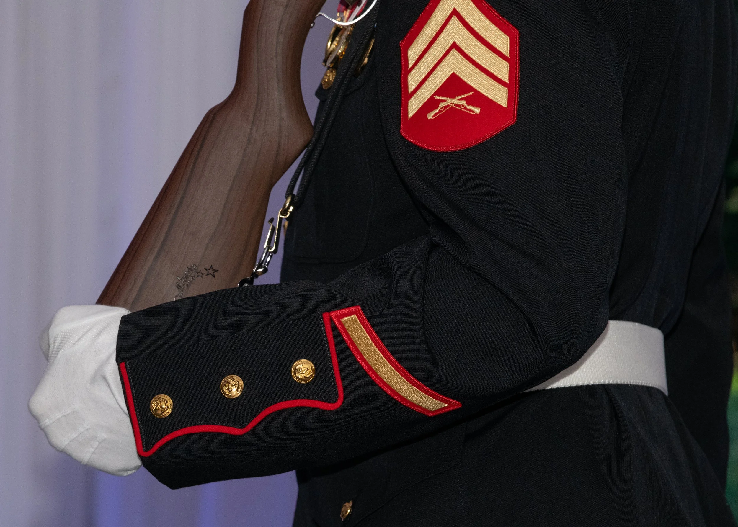 Close-up of a person dressed in a United States Marine Corps dress uniform, showing shoulder and arm details, including rank insignia, buttons, and a white glove.