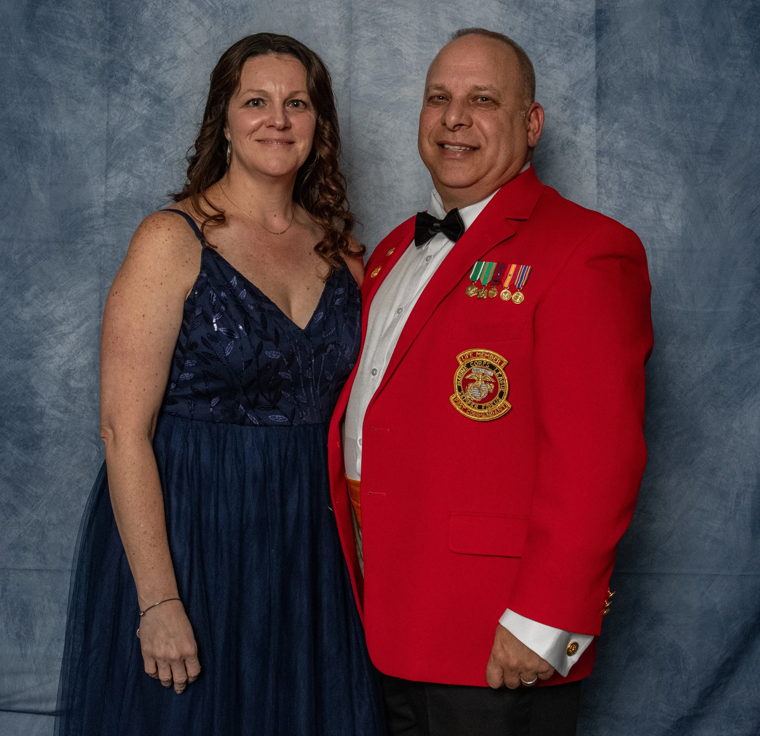 A woman in a dark blue dress and a man in a red jacket with medals pose together against a gray background.