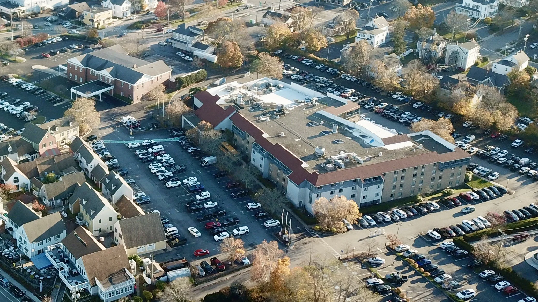 Aerial view of a residential and commercial area with a large multi-story building, numerous parking lots filled with cars, and houses, with trees showing autumn colors.