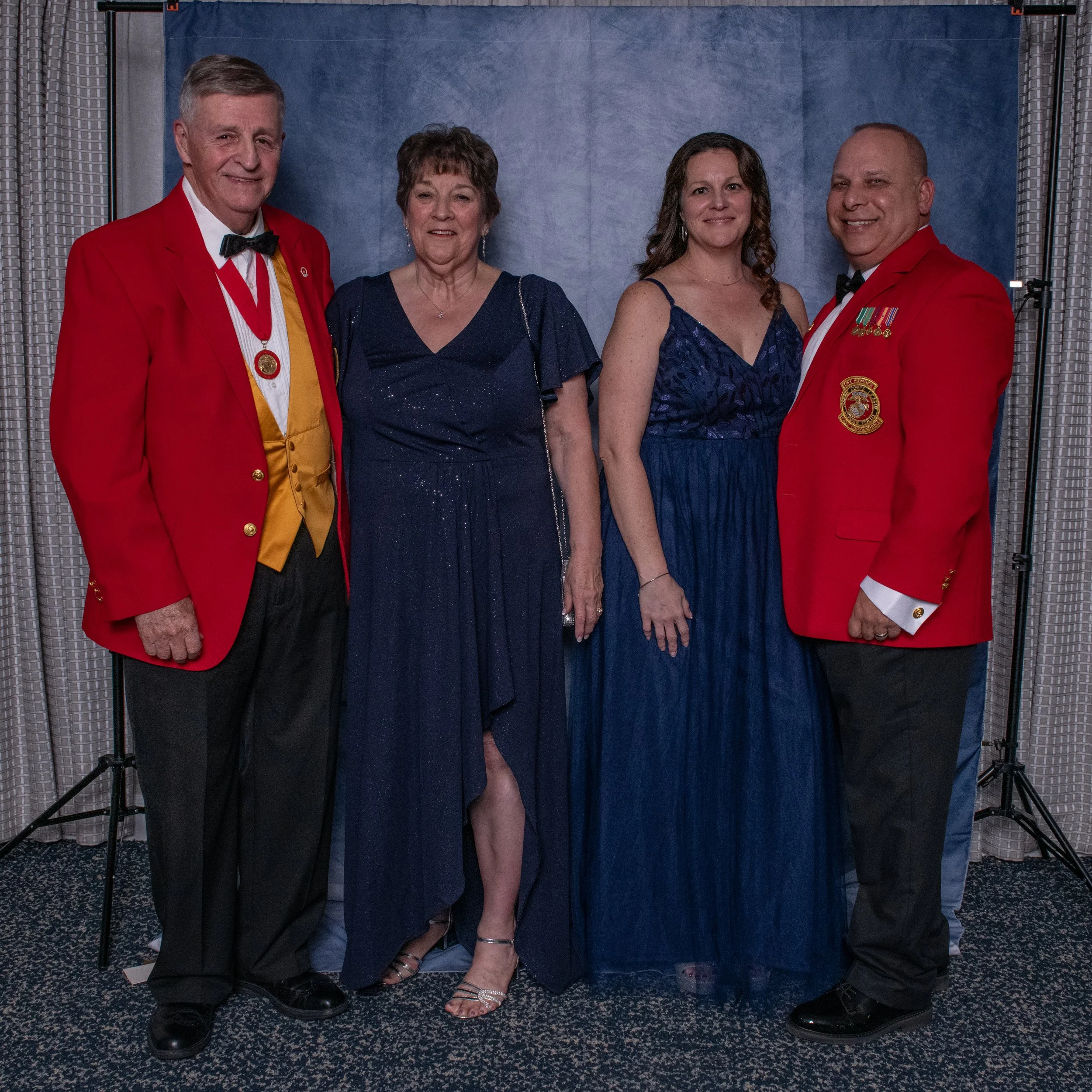 Four people standing in front of a blue backdrop at a formal event. The two men are wearing red blazers with medals, black pants, and bow ties. The women are in dark blue evening gowns. All are smiling.
