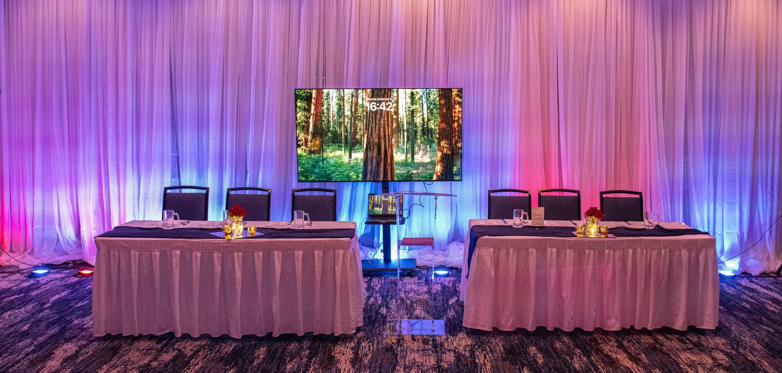Event setup with two long tables covered in white tablecloths with black runners, decorated with small vases of red roses and gold accents, against a backdrop of purple and pink lighting. A large screen displaying a forest scene is centered behind th