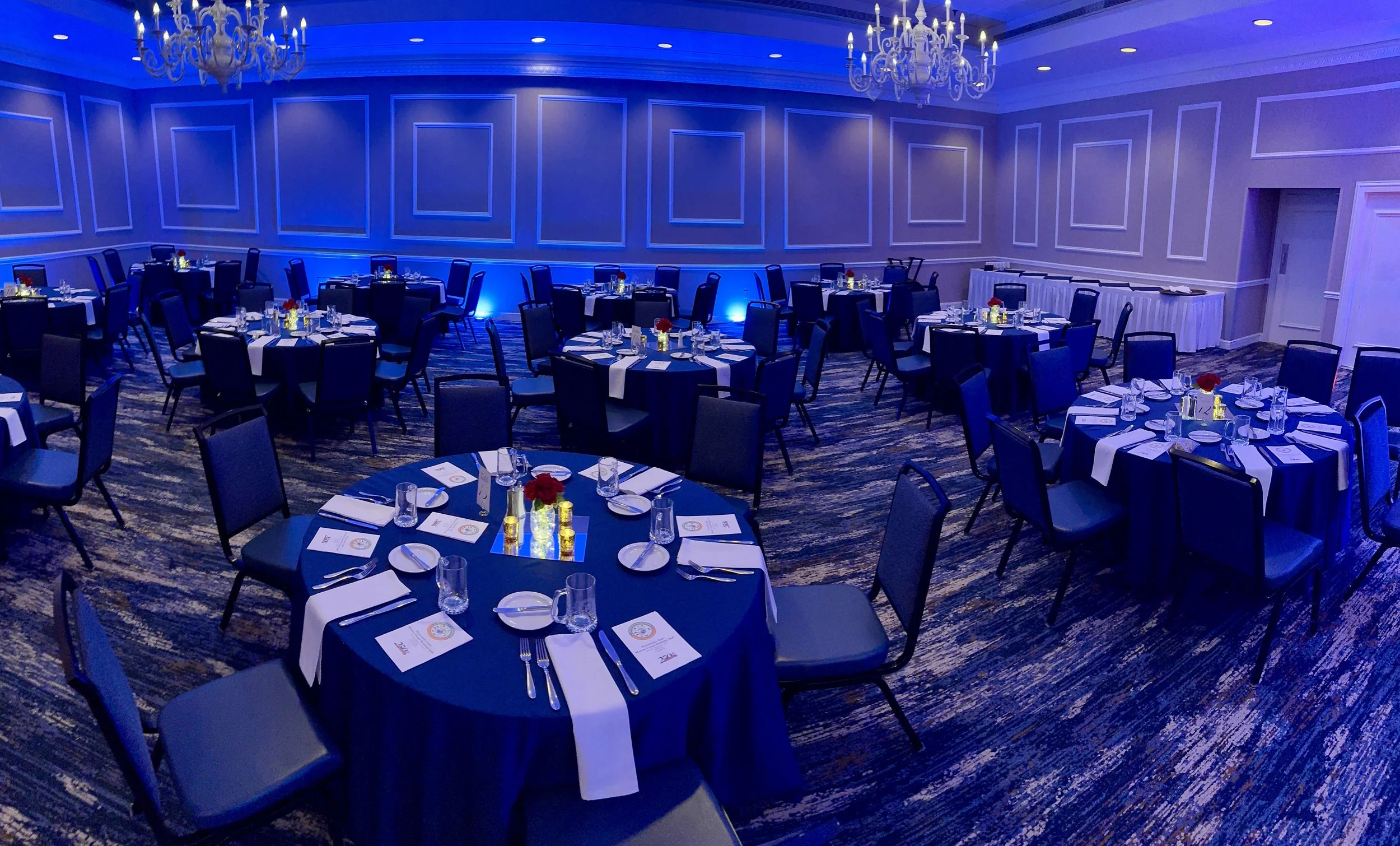 Set up banquet room with round tables adorned with navy tablecloths, white napkins, glassware, and floral centerpieces, illuminated with blue lighting and chandeliers.