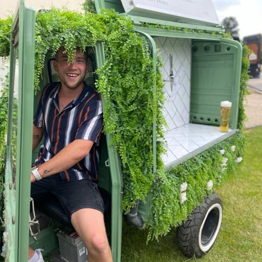 A smiling young man with a striped shirt sitting inside a small, green prosecco trailer decorated with green plants and white flowers, with a large glass of beer on the counter.