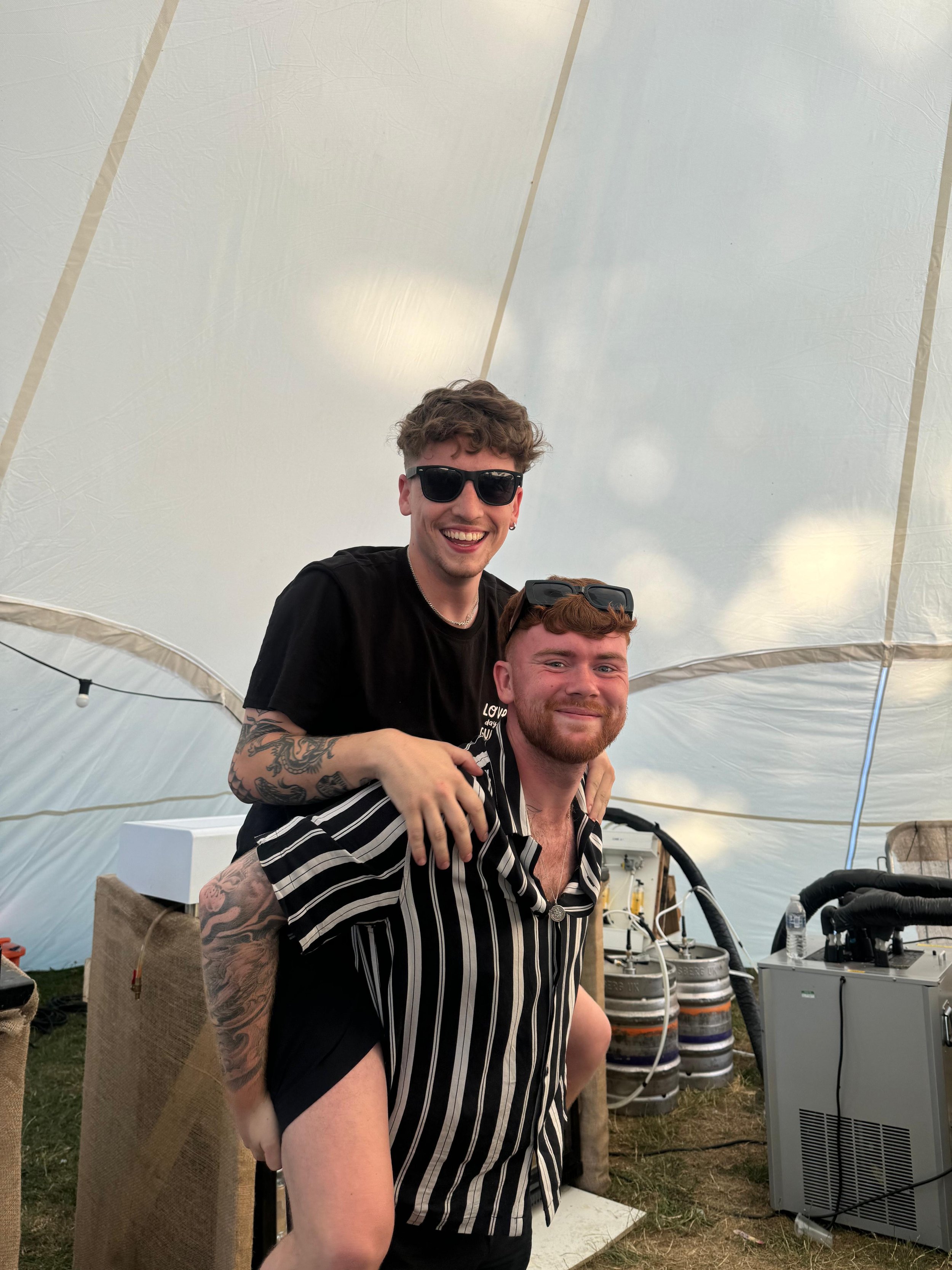 Two young men smiling indoors, one giving the other a piggyback ride. Both are wearing sunglasses, and the background shows a tent with some equipment and beer kegs.