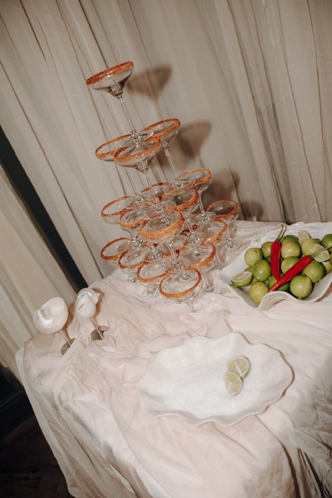 A pyramid of empty cocktail glasses with salted rims, a plate with two lime wedges, and a bowl of green apples, red chili peppers, and limes on a table covered with a light-colored cloth.