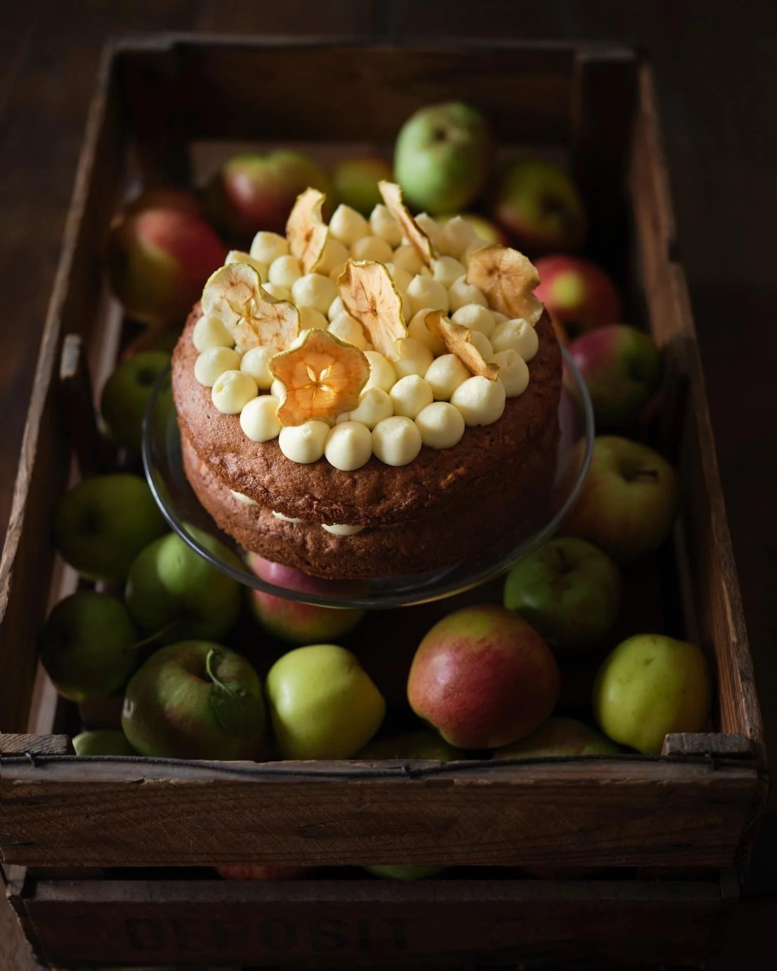 Chocolate cake topped with white cream, apple chips, and white chocolate balls, placed on a glass plate inside a wooden crate filled with apples.