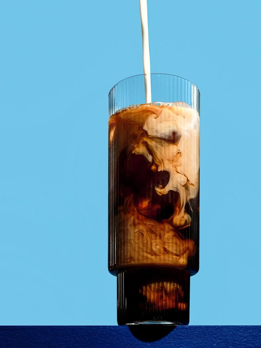 A tall glass with vertical ridges being filled with a dark beverage, likely iced coffee, as white milk swirls into it against a blue background.