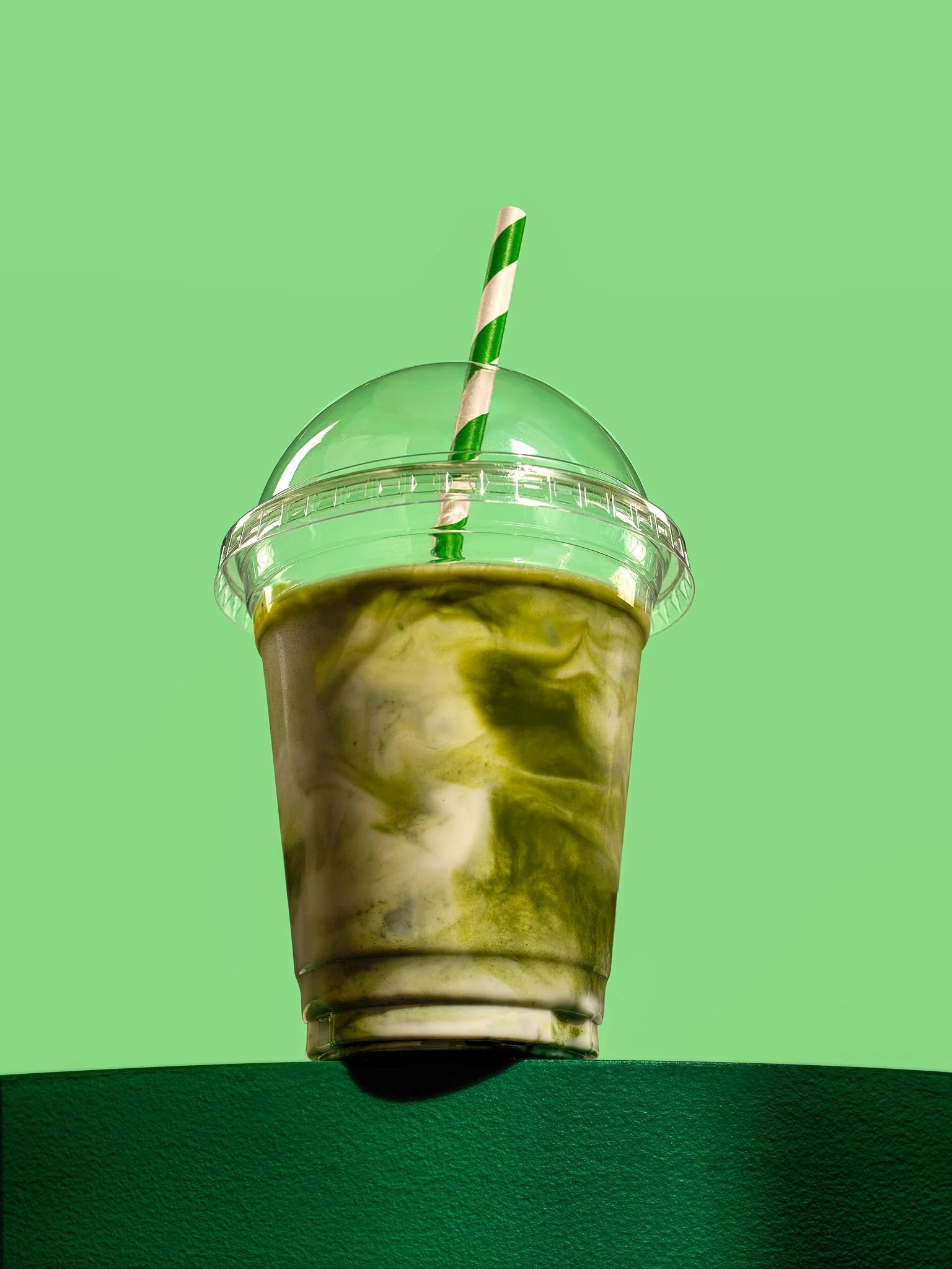 Green and white marble iced beverage in a clear plastic cup with a dome lid and green and white striped straw against a green background.