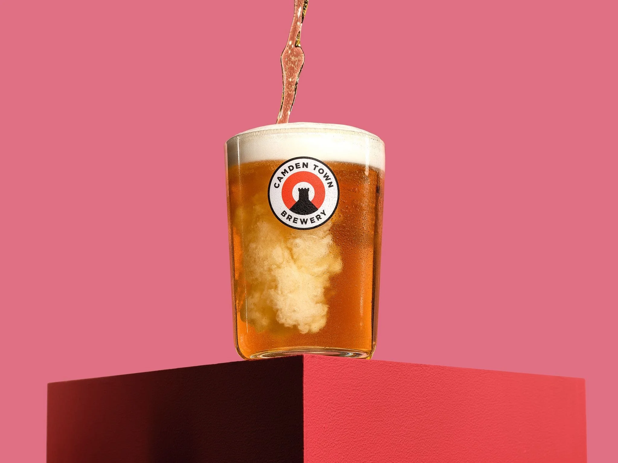 A glass of beer with foam on top, placed on a red surface against a pink background, with a hand pouring more beer into the glass.