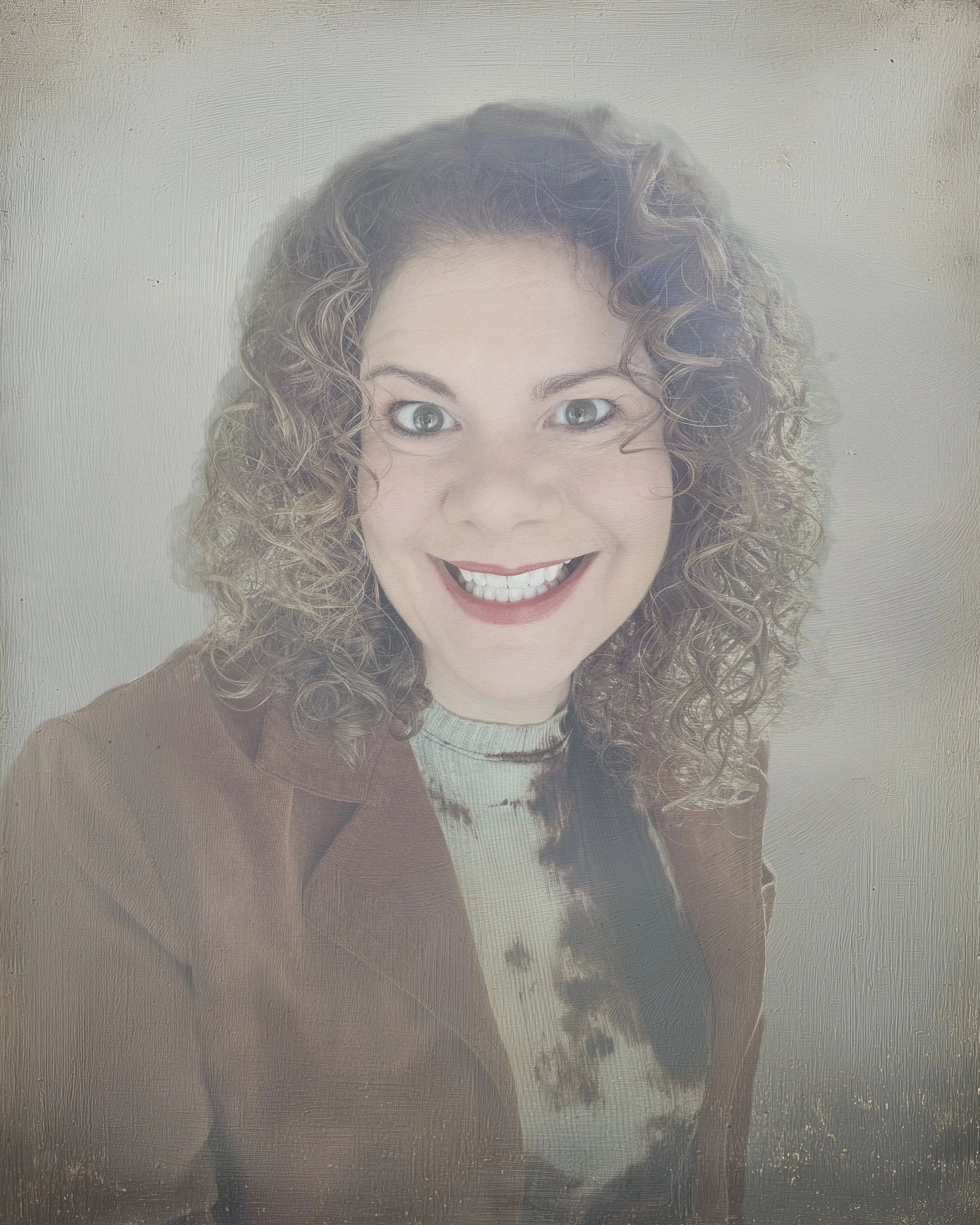 A woman with curly hair smiling while looking at the camera, wearing a brown jacket and a tie-dye shirt, standing in front of a plain textured wall.