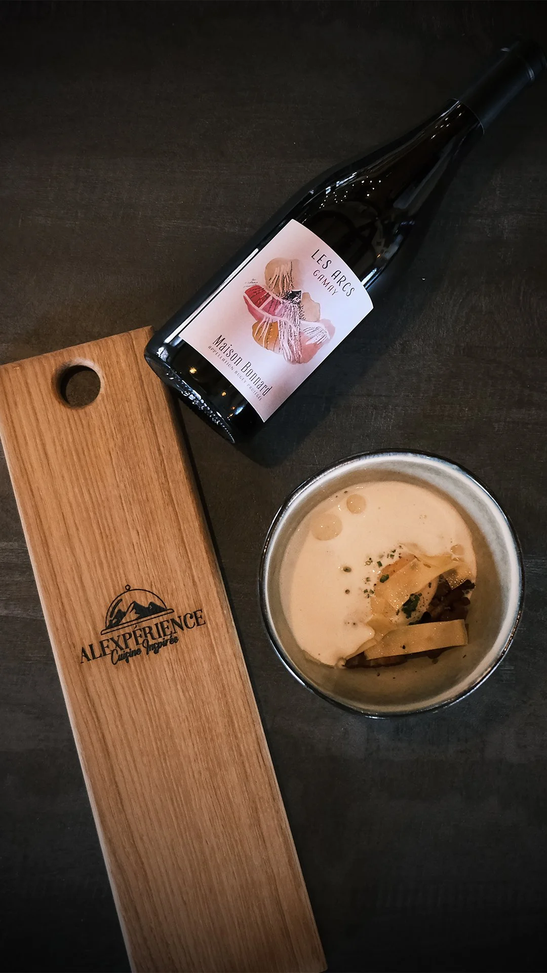Une bouteille de vin rouge du domaine Bonnard en Savoie, une entrée oeuf parfait et beaufort, et une planche en bois avec le logo "Alexperience Cuisine Inspirée".
