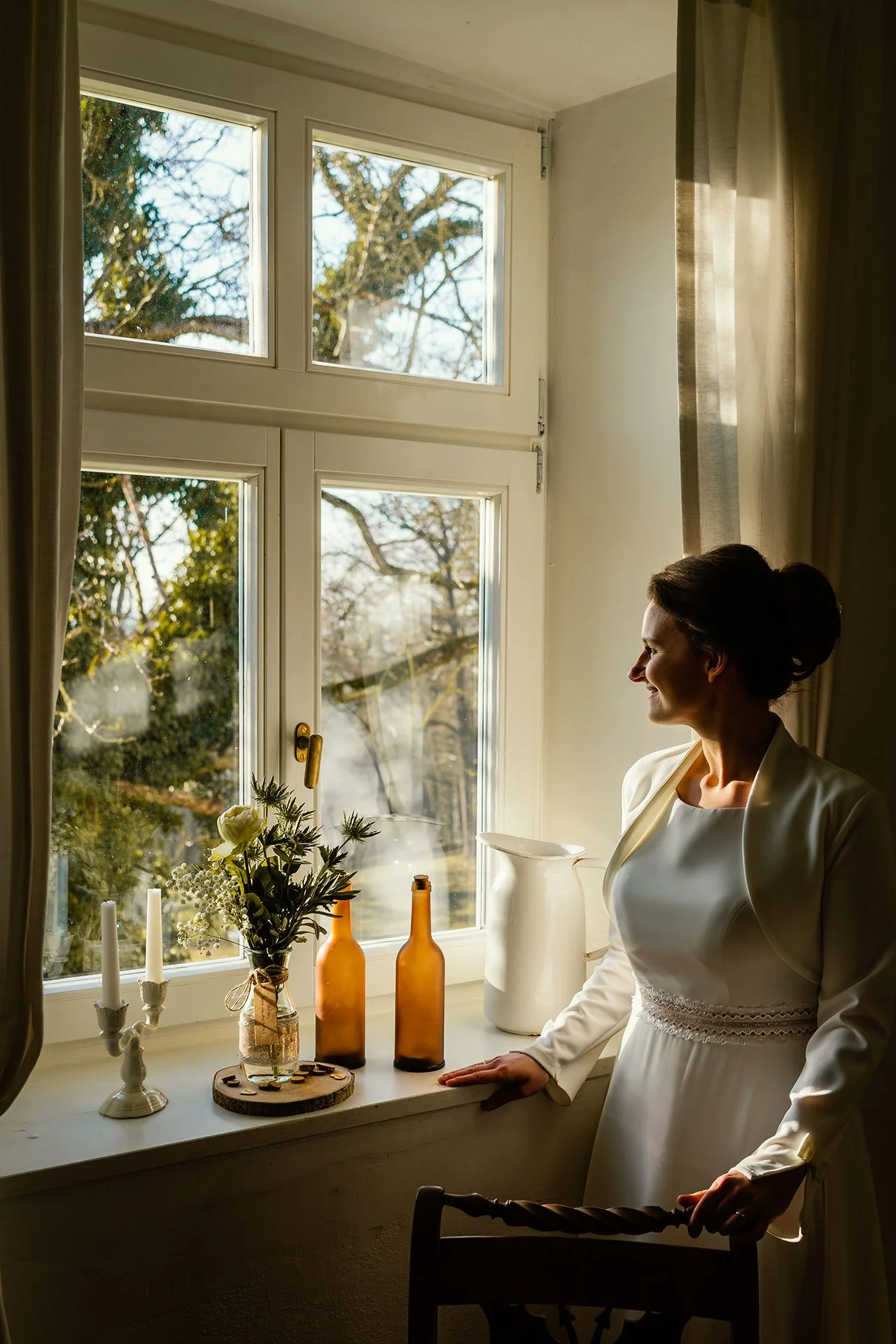 Frau in weißem Kleid steht am Fenster und schaut nach draußen, mit Sonnenlicht auf ihrem Gesicht. Fenster mit Blick auf Bäume, auf der Fensterbank dekorative Elemente wie Blume, zwei Flaschen und Kerzenständer.