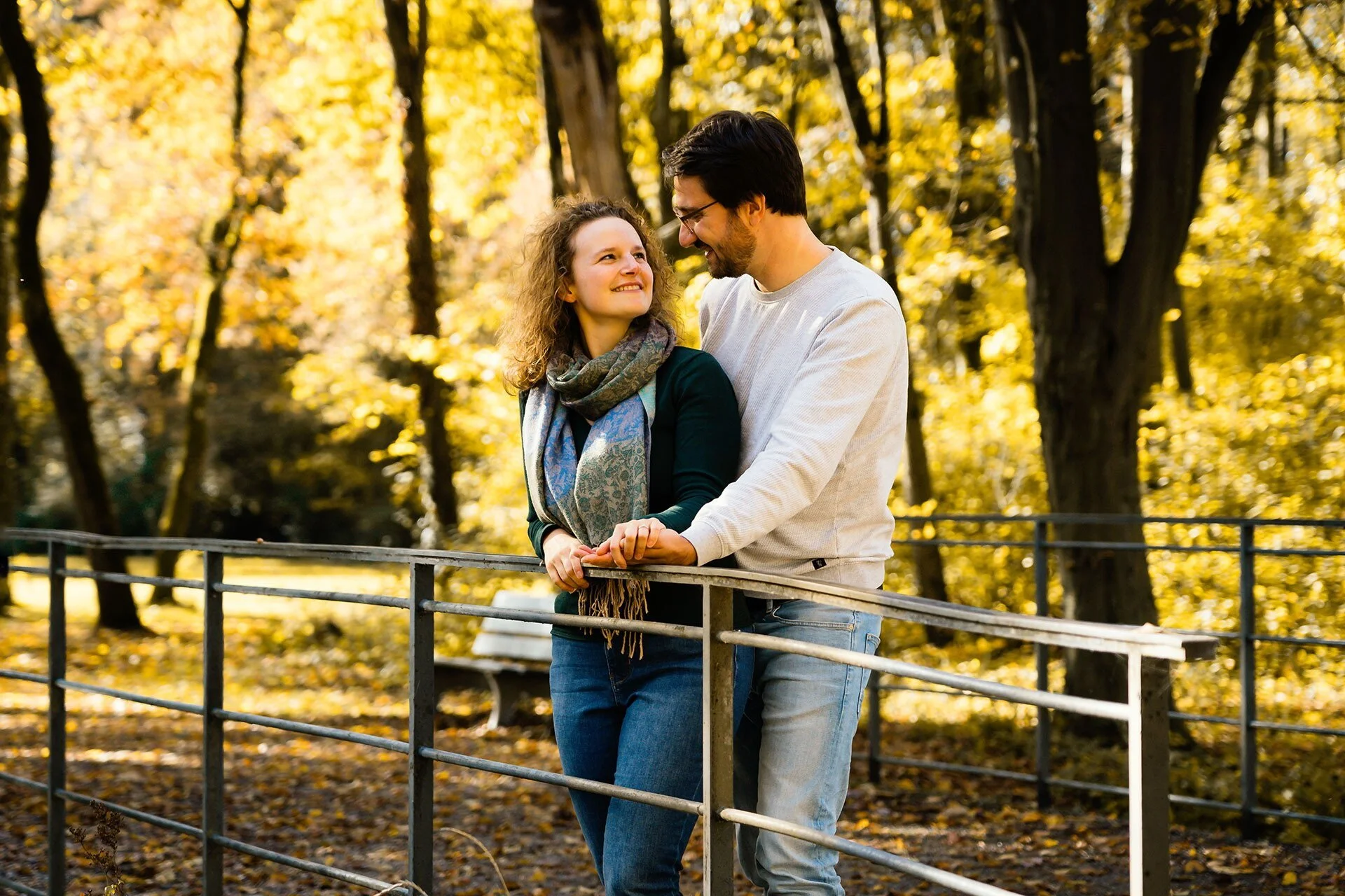 Ein glückliches Paar steht im Herbst im Park, hält sich an den Händen und schaut sich liebevoll an, umgeben von goldfarbenen Blätter und schwarzen Baumstämmen.