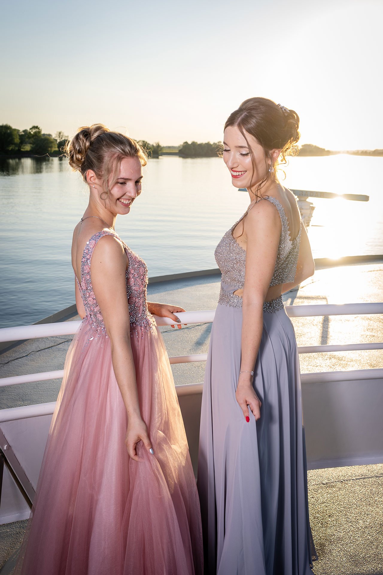 Zwei Frauen in eleganten Abendkleidern stehen auf einem Boot neben einem Fluss bei Sonnenuntergang, lächeln und schauen sich glücklich an.