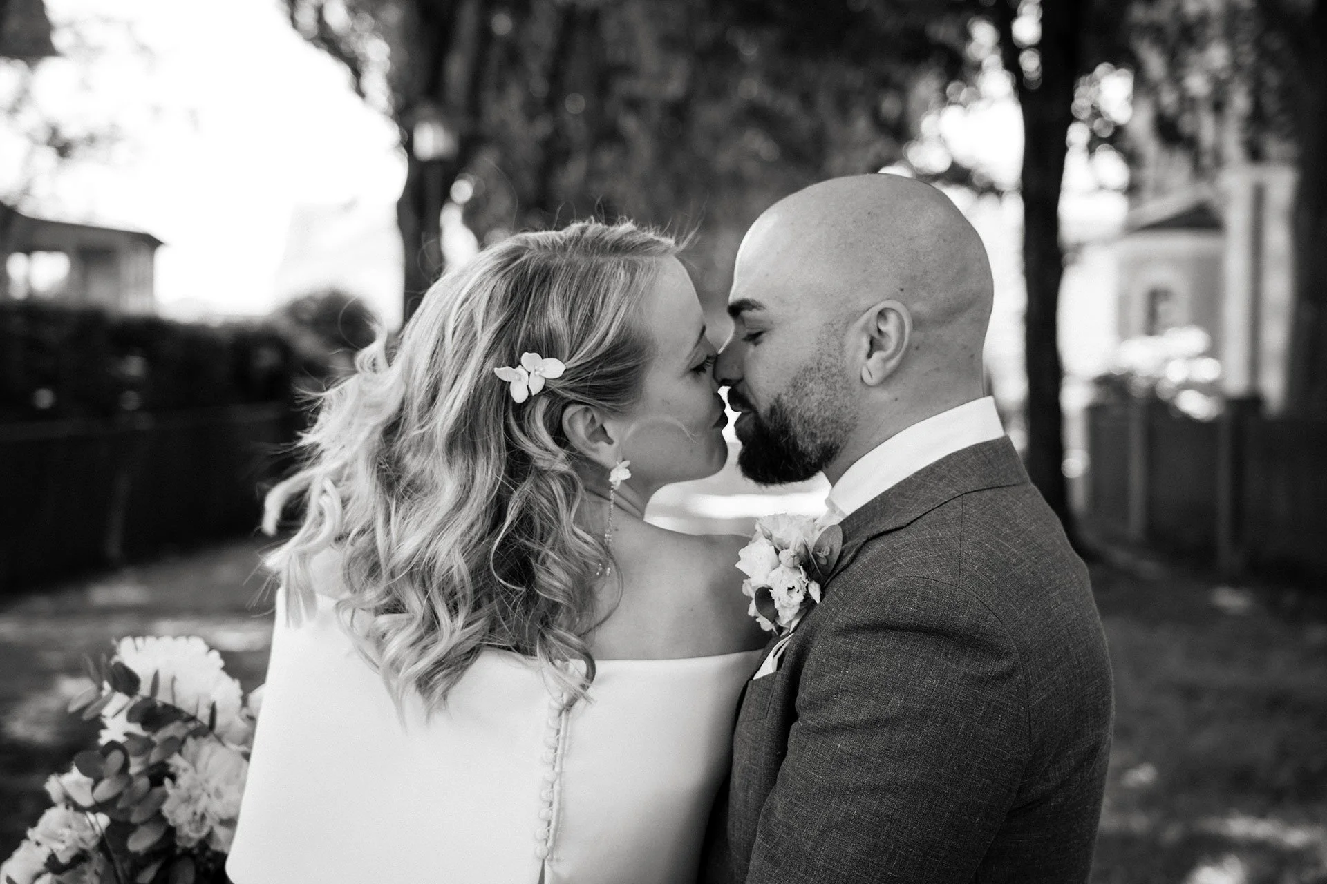 Braut und Bräutigam stehen sich bei einer Hochzeit draußen sehr nah, die Stirn an Stirn, mit sichtbaren Blumen im Haar der Braut und im Anzug des Bräutigams, umgeben von Bäumen und Gebäuden im Hintergrund, schwarz-weiß Aufnahme.