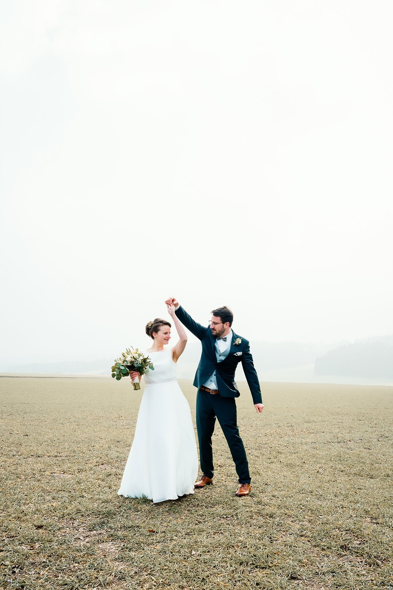 Ein Brautpaar tanzt im offenen Feld an einem grauen Tag, die Braut trägt ein weißes Kleid und hält einen Blumenstrauß, während der Bräutigam in einem dunklen Anzug mit Brille zu sehen ist.