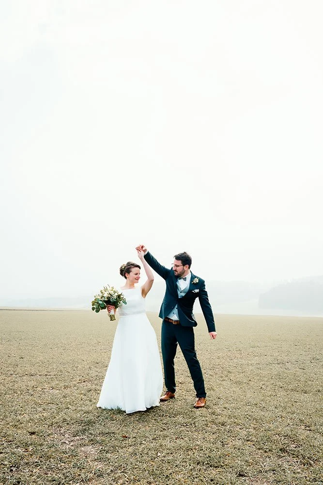 Ein Brautpaar tanzt auf einer offenen Wiese bei einem Hochzeitsfoto, die Braut trägt ein weißes Kleid und hält einen Blumenstrauß, der Bräutigam trägt einen dunklen Anzug.