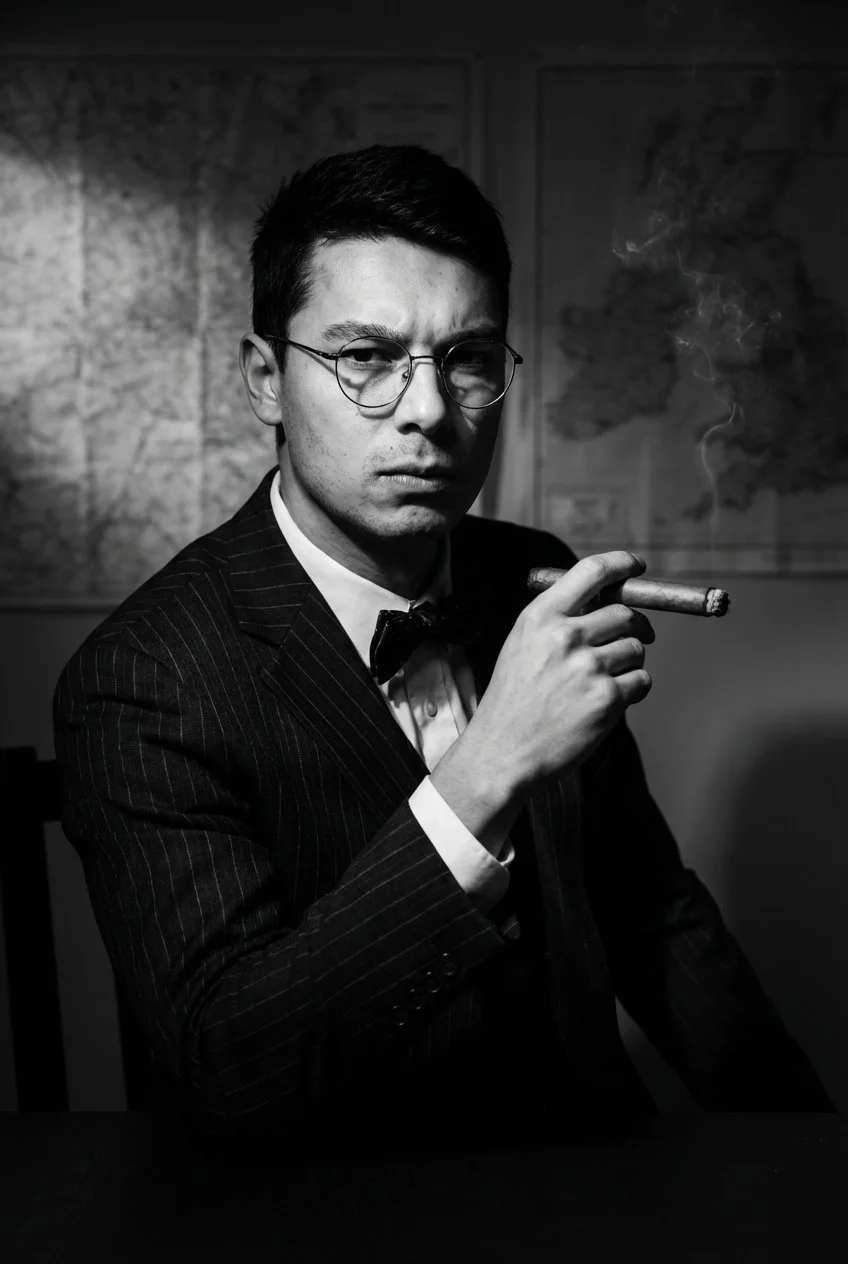 Ein junger Mann in einem schwarzen Anzug mit weißem Hemd und schwarzer Fliege sitzt an einem Tisch, hält eine Zigarre in der Hand und schaut ernst in die Kamera. Im Hintergrund sind Landkarten an einer Wand zu sehen.
