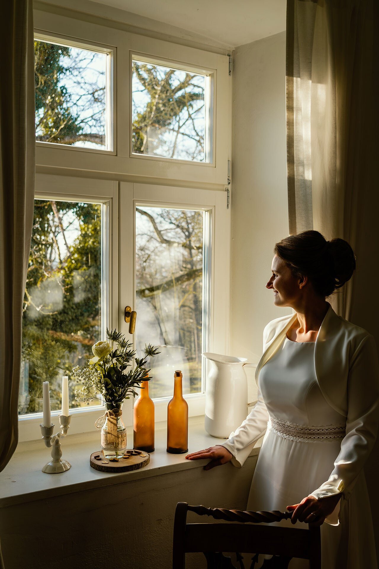 Eine Frau in einem weißen Kleid steht neben einem Fenster, durch das Sonnenlicht scheint. Auf dem Fensterbrett stehen zwei braune Flaschen, eine weiße Keramikvase, ein Blumenstrauß und Kerzen. Draußen sind Bäume mit herbstlichen Blättern zu sehen.