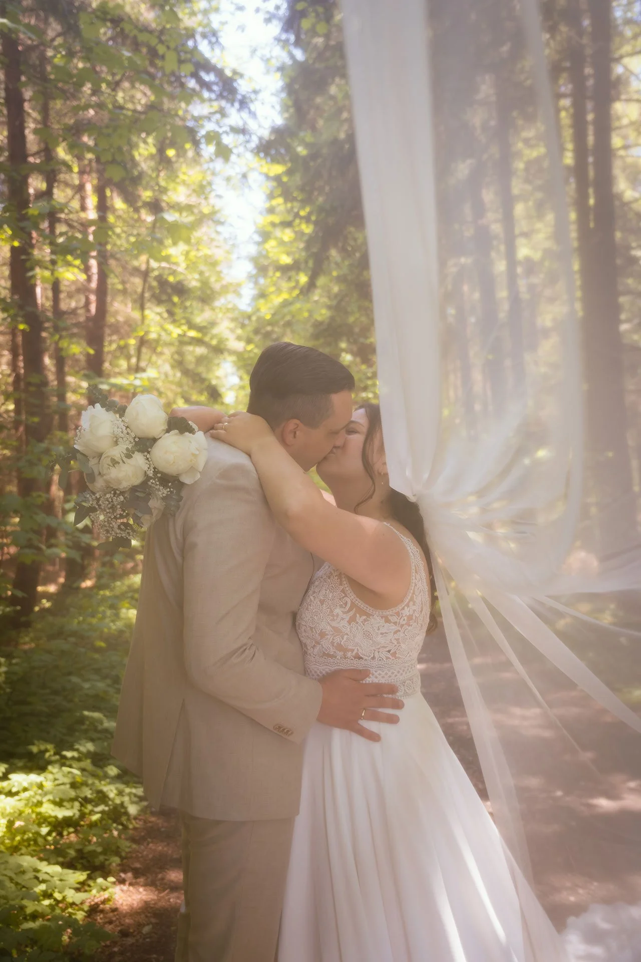 Ein Brautpaar küsst sich in einem Wald, die Braut trägt ein weißes Brautkleid mit Spitze, der Bräutigam trägt einen hellen Anzug, im Hintergrund sind Bäume und Sonnenlicht.