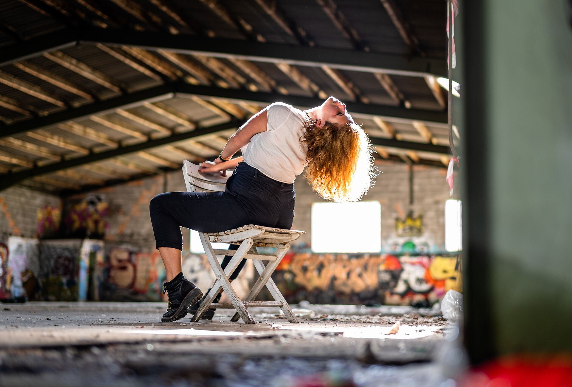 Eine junge Frau mit roten Locken sitzt auf einem alten Holzstuhl in einer abenteuerlich wirkenden Fabrikanlage, lehnt ihren Kopf nach hinten, während sie sich entspannt. Die Umgebung ist industrial, mit grauen Ziegelwänden, Graffiti auf der Wand im Hintergrund und einem dunklen, gewölbten Dach.