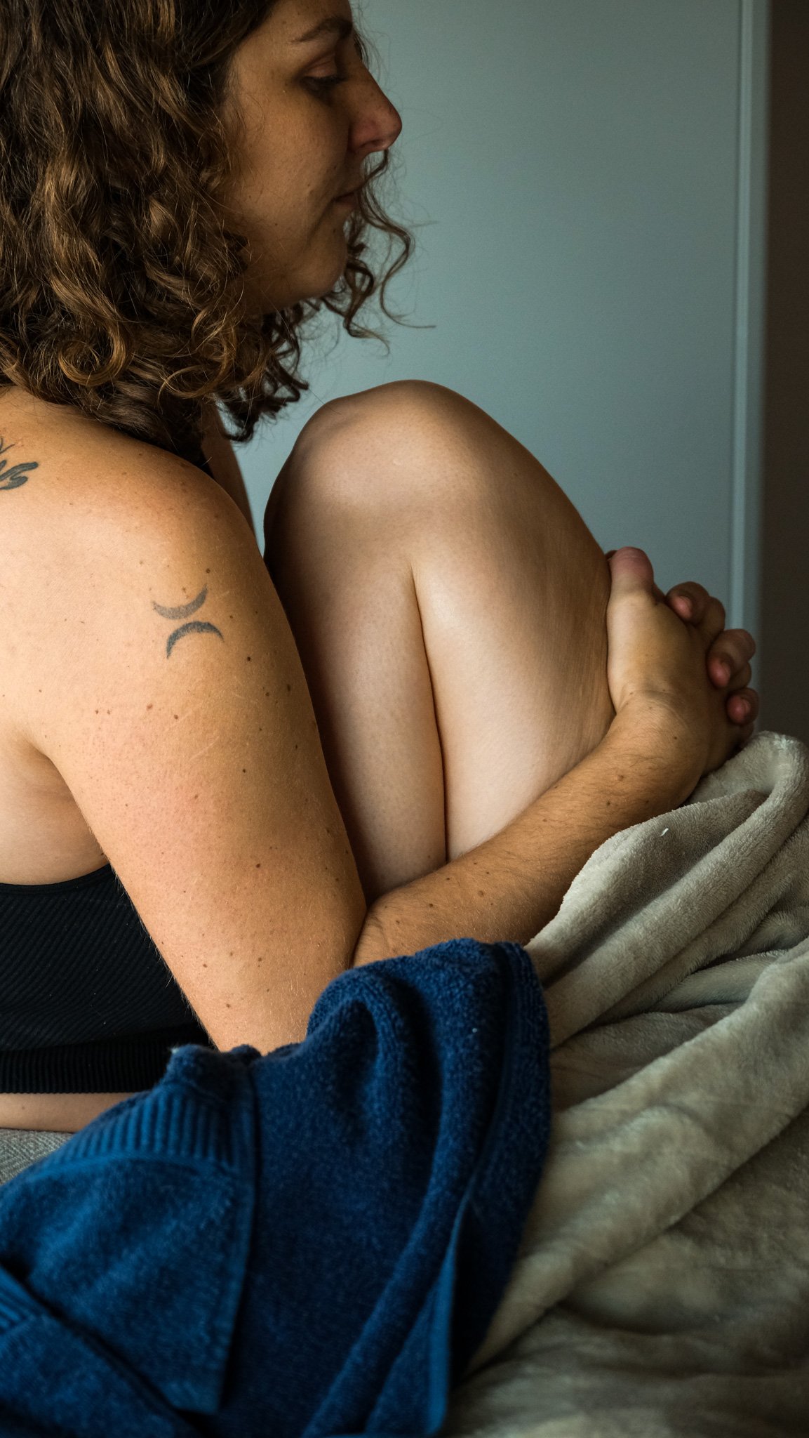 Une femme assise, tenant une jambe pliée contre sa poitrine, vêtue d'un haut noir, dans une pièce avec mur blanc.