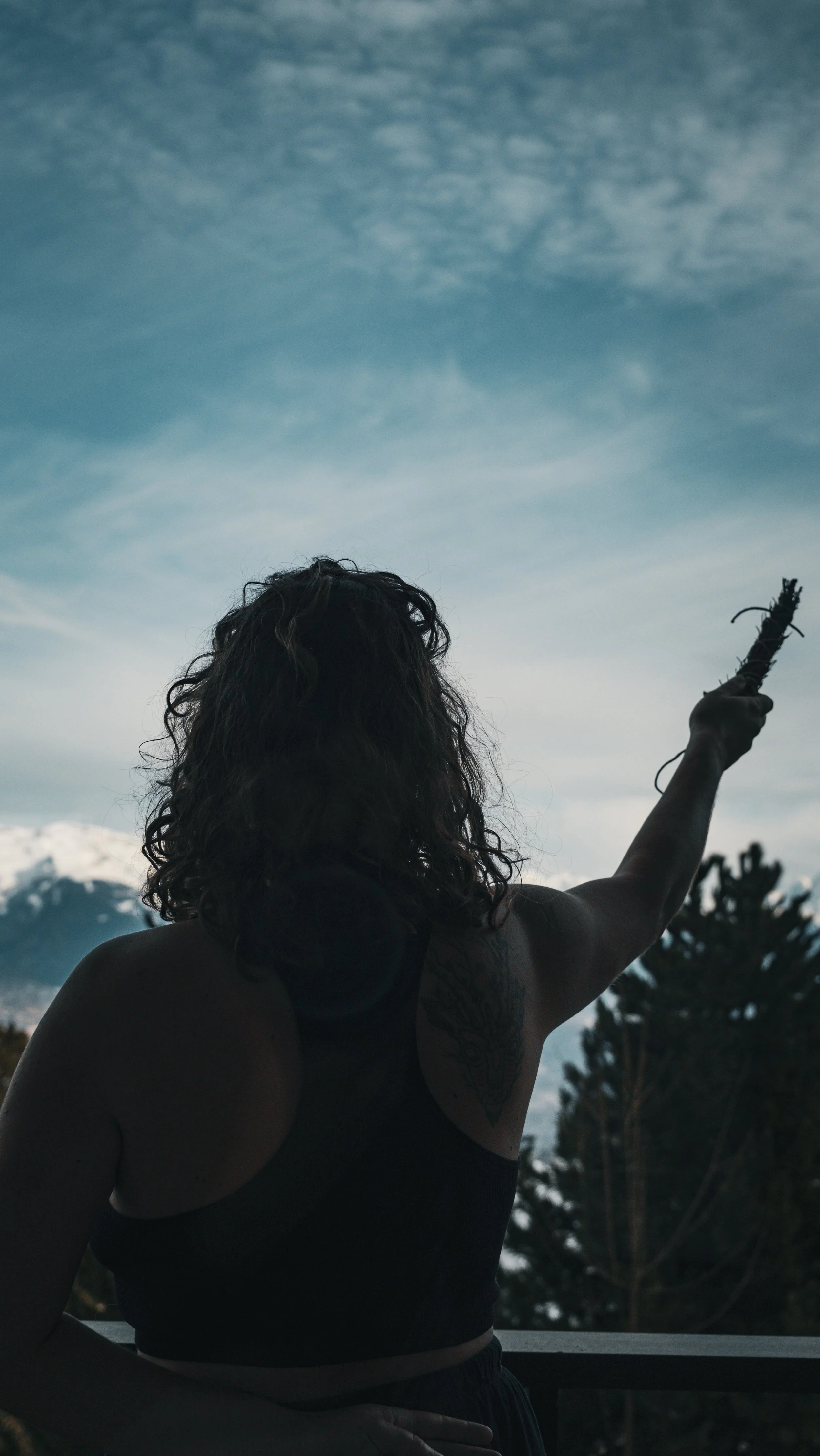 Une femme avec des cheveux bouclés, portant un débardeur noir, vue de dos, tenant un bâton de fumigation  levé vers le ciel, avec des arbres et des nuages en arrière-plan.