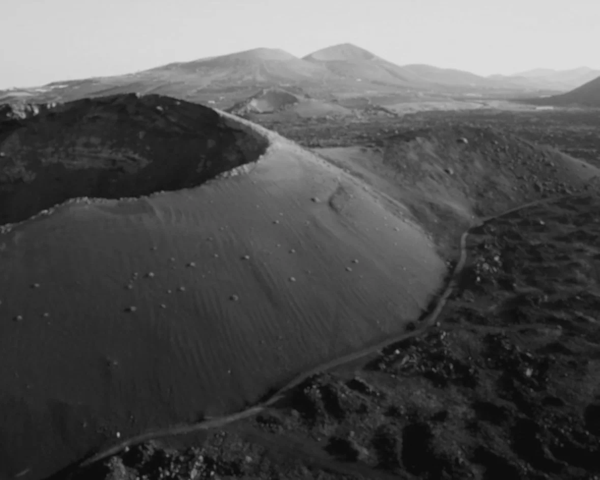 Black and white photo of a volcanic mountain range with rugged terrain, hills, and a trail winding through the landscape.