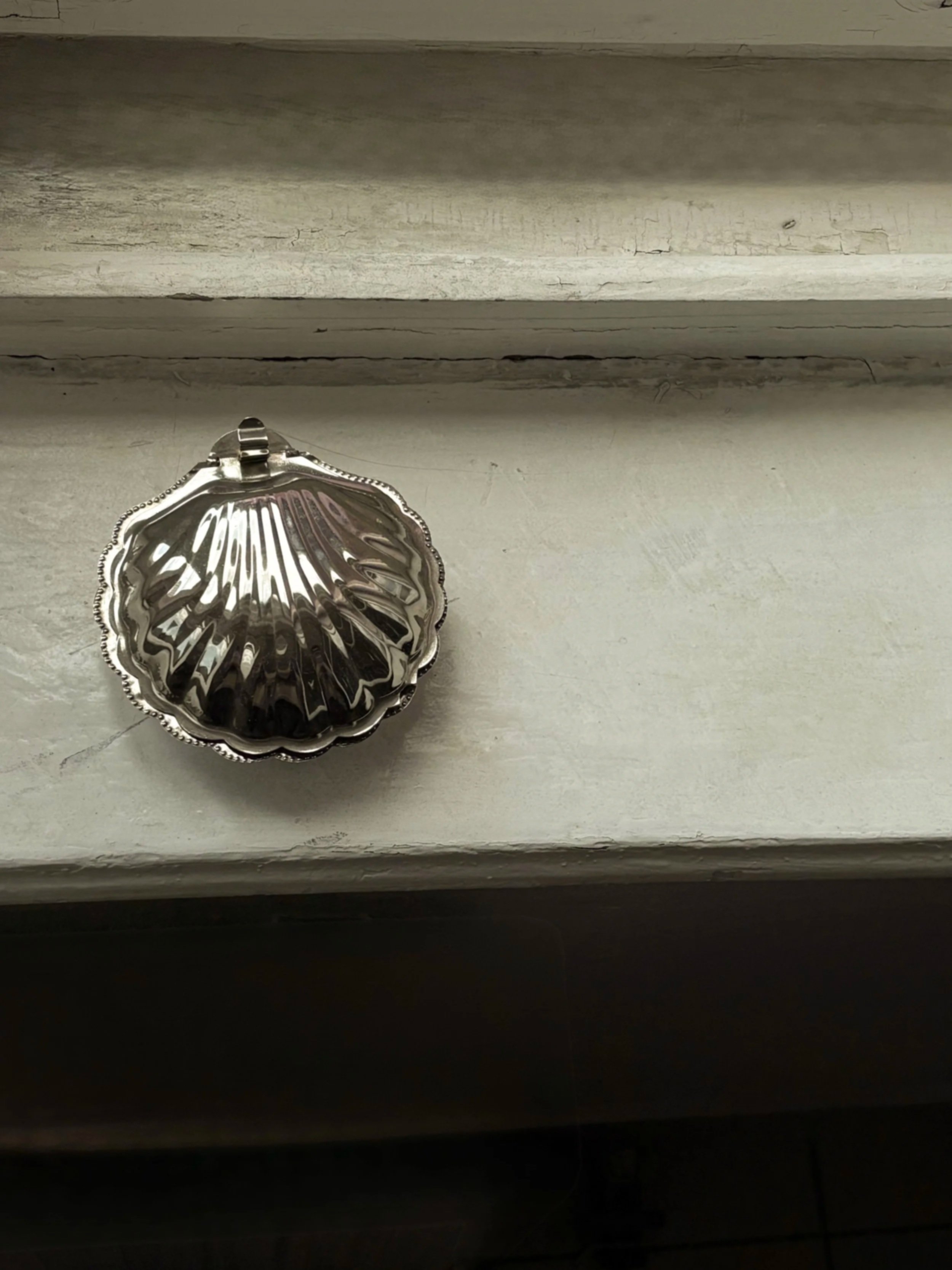 A silver seashell-shaped pendant with detailed ridges, resting on a white windowsill.