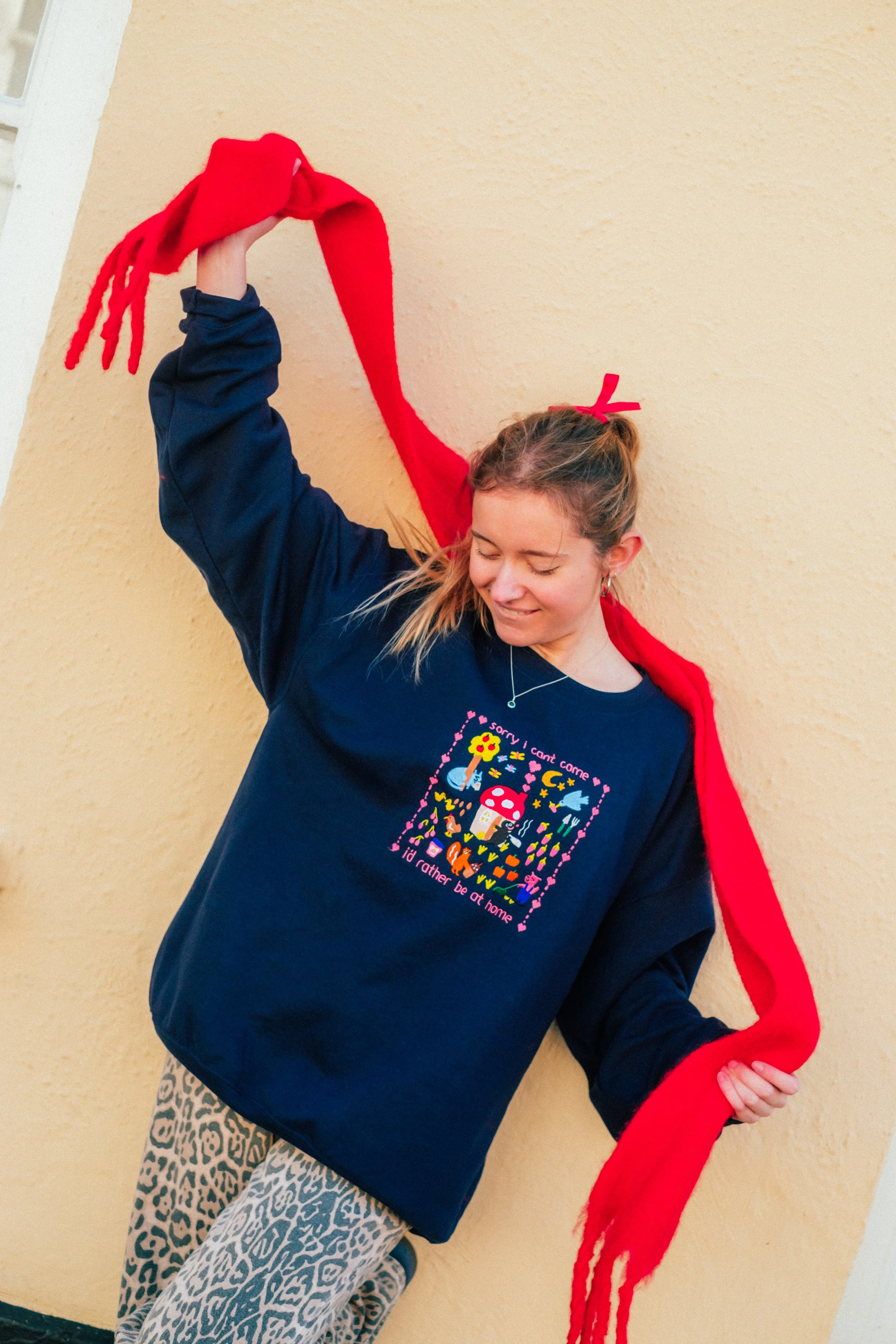 Young woman wearing a navy sweatshirt with colorful pixel art and text, holding a red scarf, and standing against a beige textured wall.