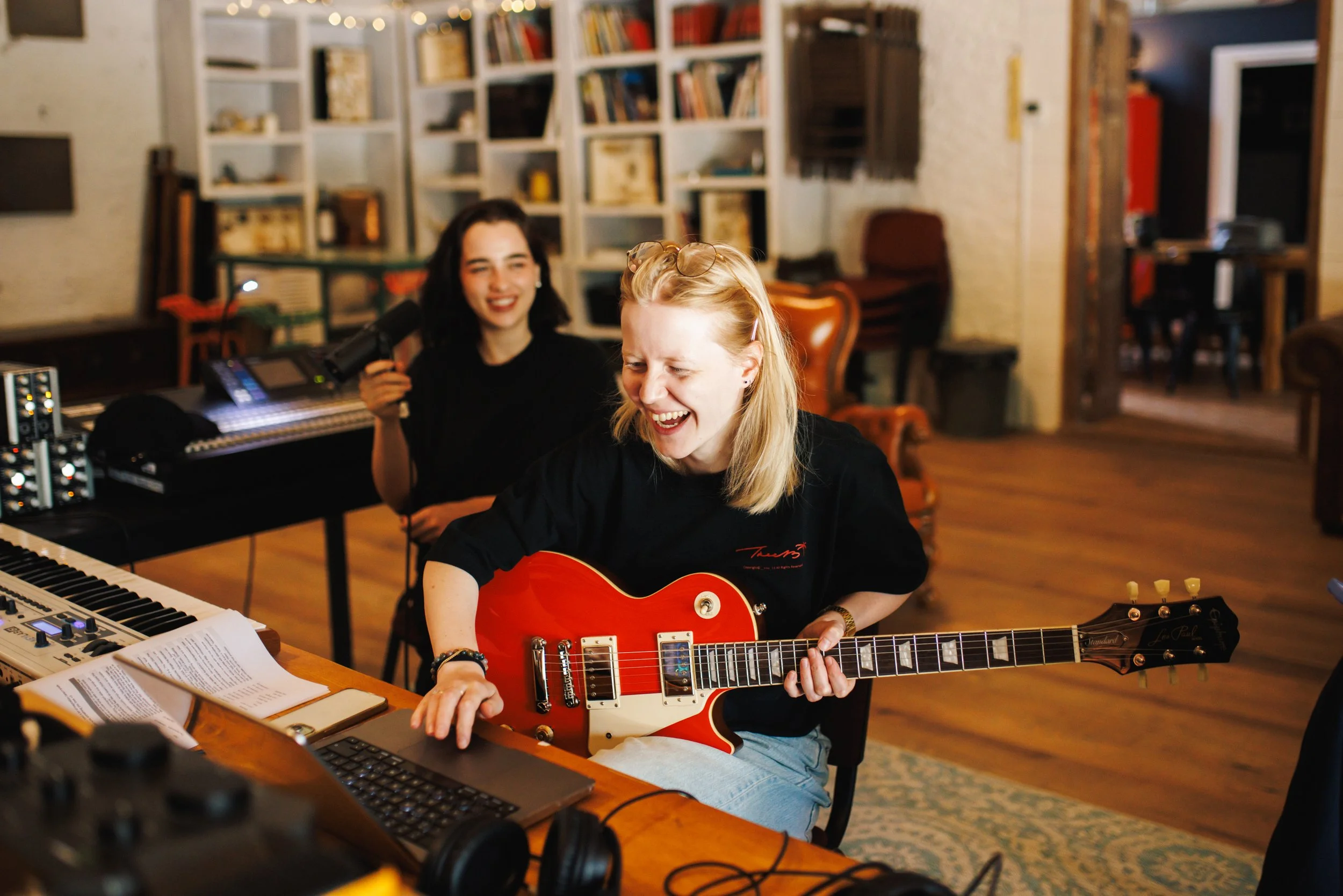 Two women in a music studio, one playing an electric guitar and the other holding a microphone, surrounded by musical equipment and books, both smiling and enjoying the moment.
