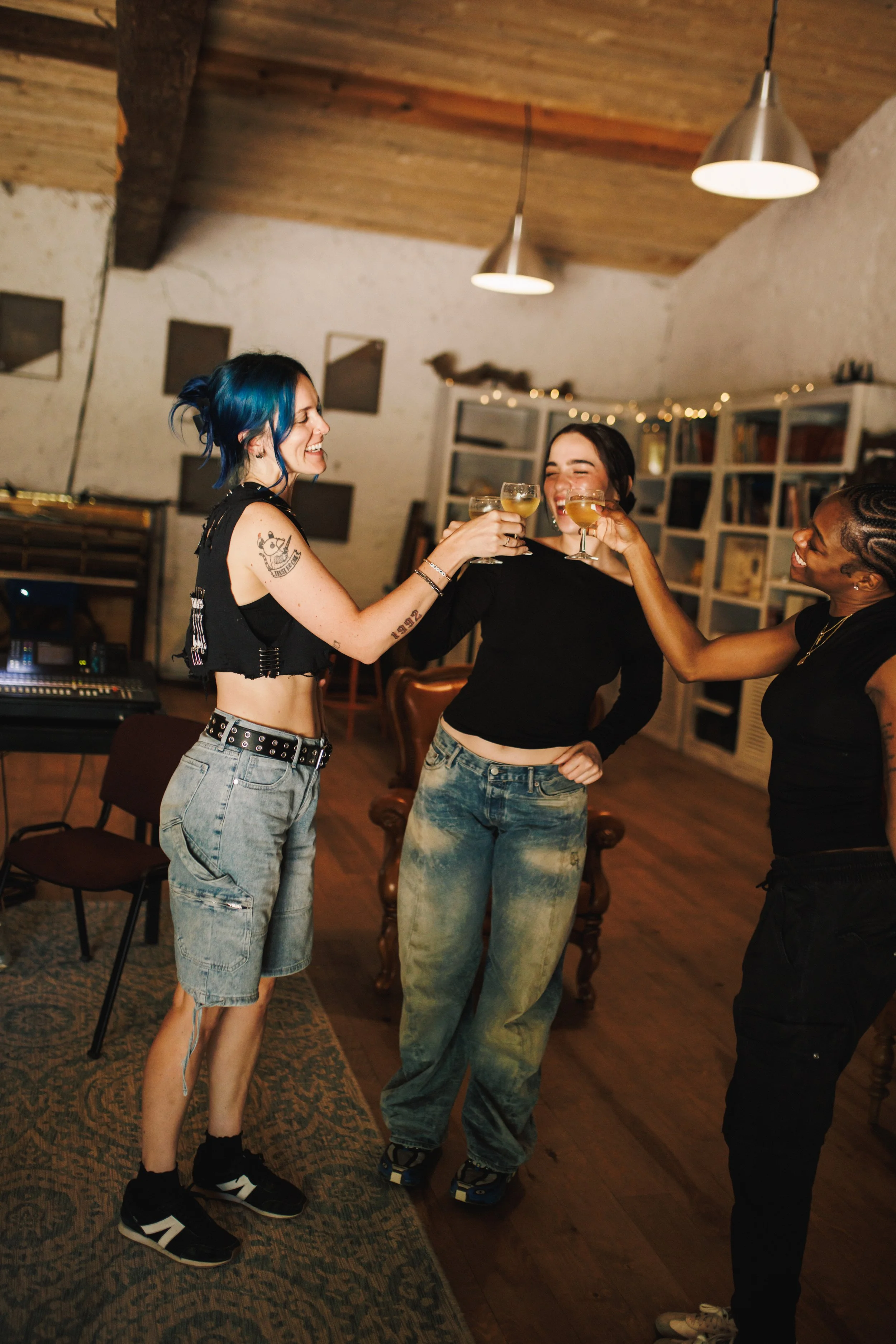 Three women celebrating together indoors, raising their glasses for a toast, smiling, with warm lighting and a cozy background.