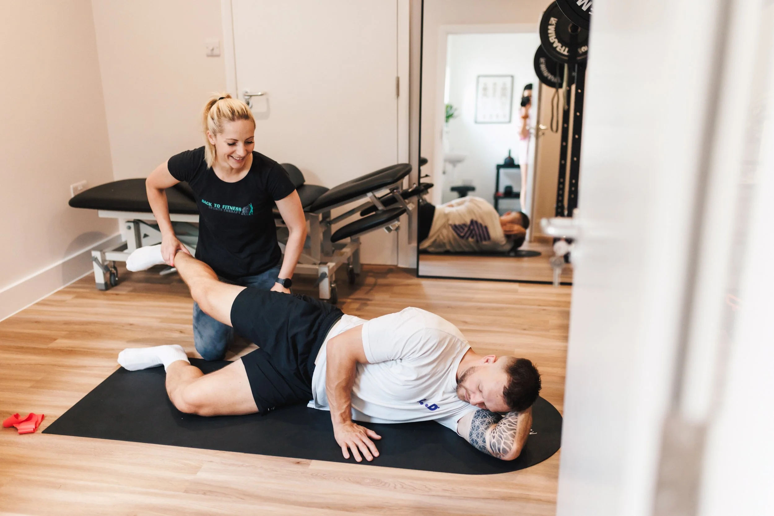 A physical therapist assisting a man with a leg stretch on a mat in a fitness or therapy room, with exercise equipment and a mirror in the background.