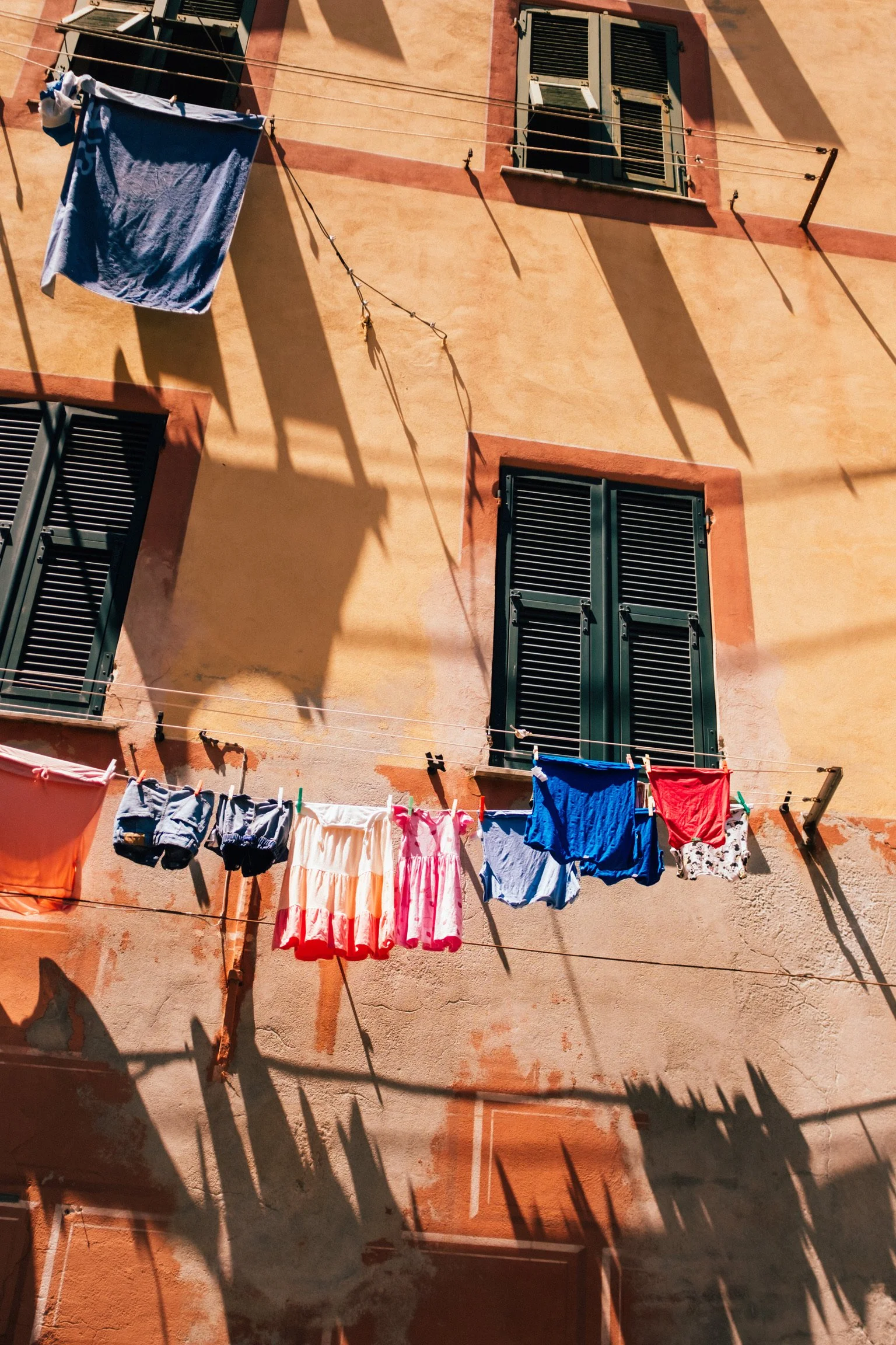 Clothes hanging on clotheslines outside a peach-colored building with green shutters, with shadows cast on the wall from the clothes and lines.