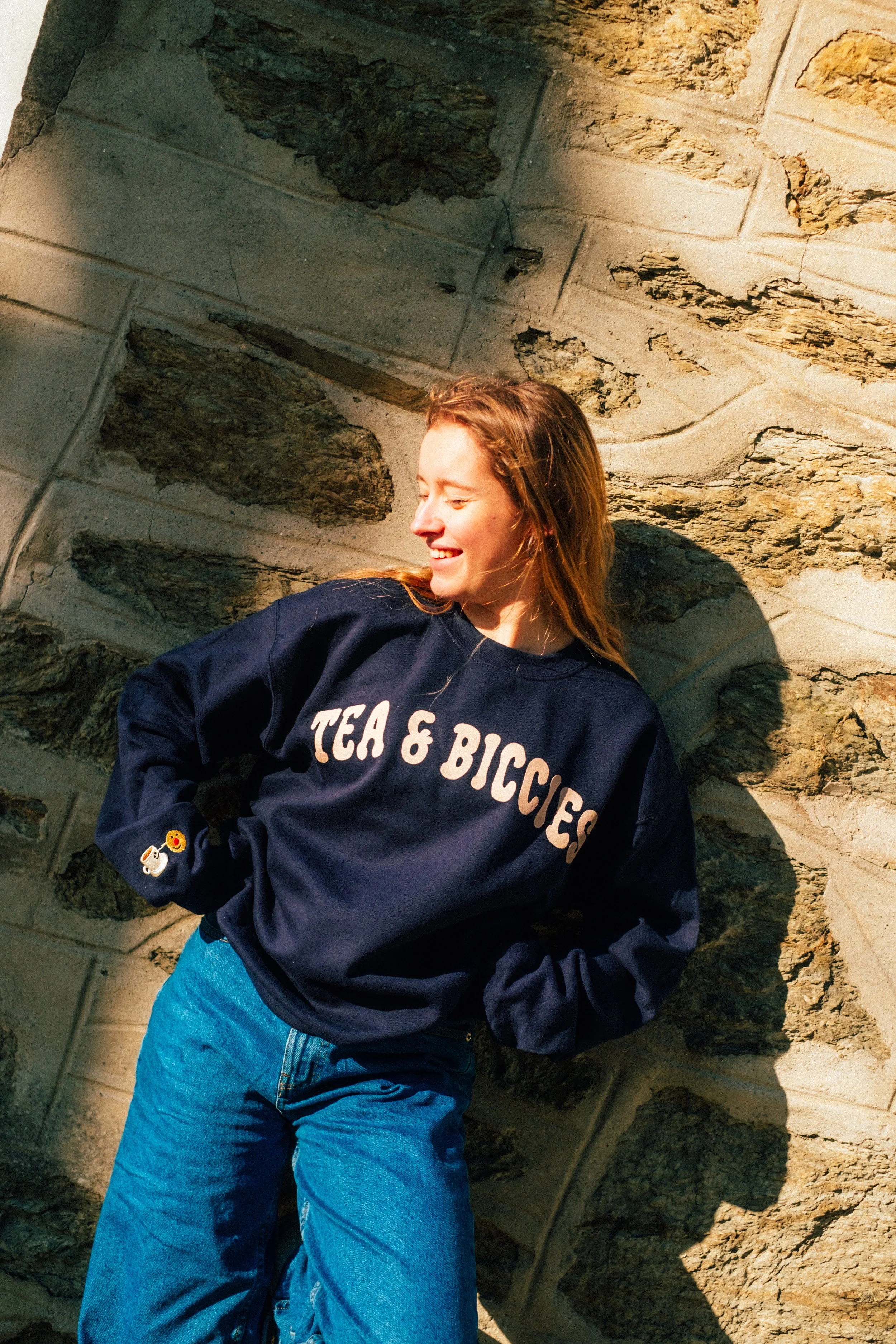 Young woman with reddish hair smiling and posing against a stone wall, wearing a navy sweatshirt with "TEA & BICCIES" written on it and jeans.
