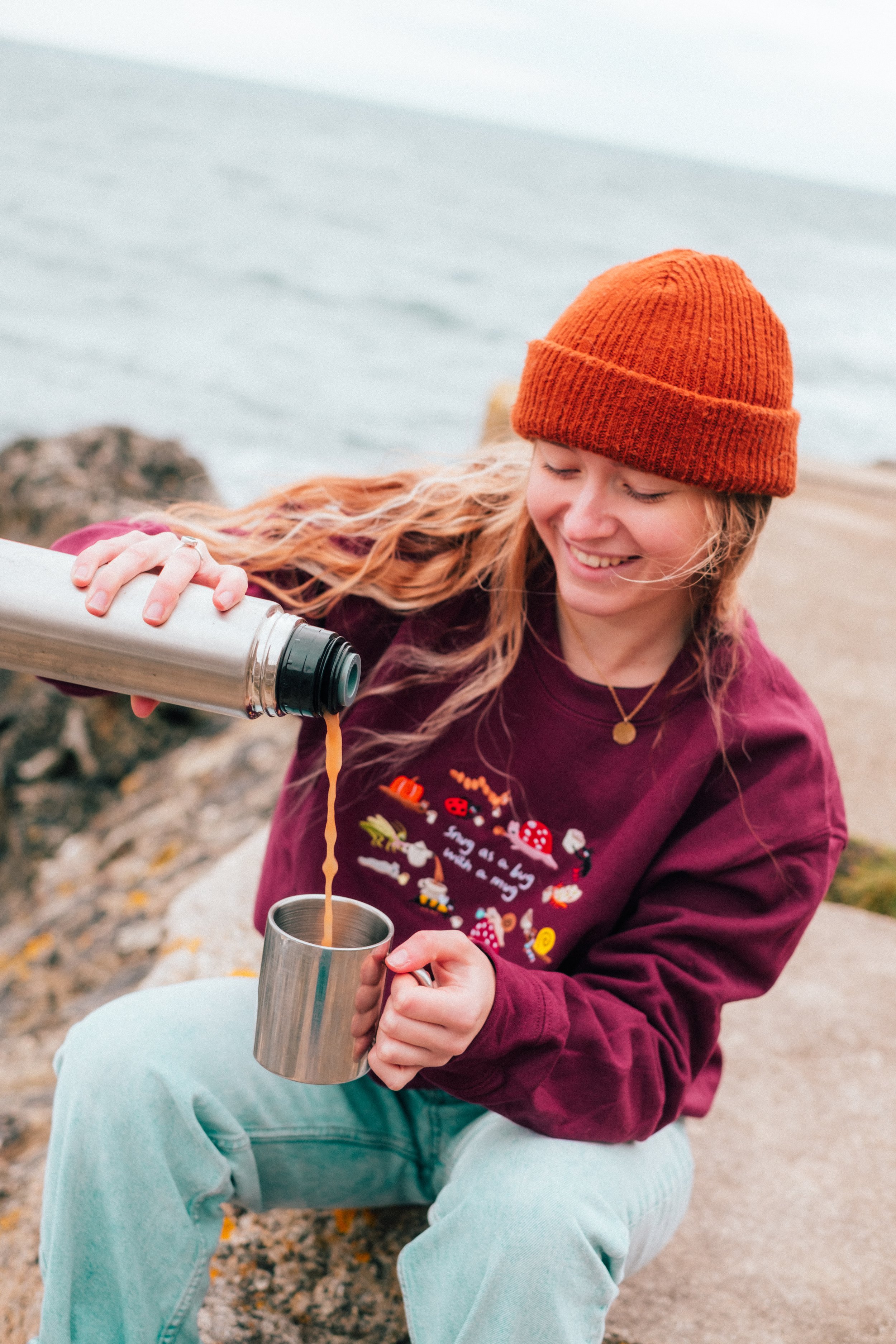 A young woman with long blonde hair wearing an orange beanie and a maroon sweatshirt sitting outdoors near water, pouring a hot beverage into a metal mug, smiling.