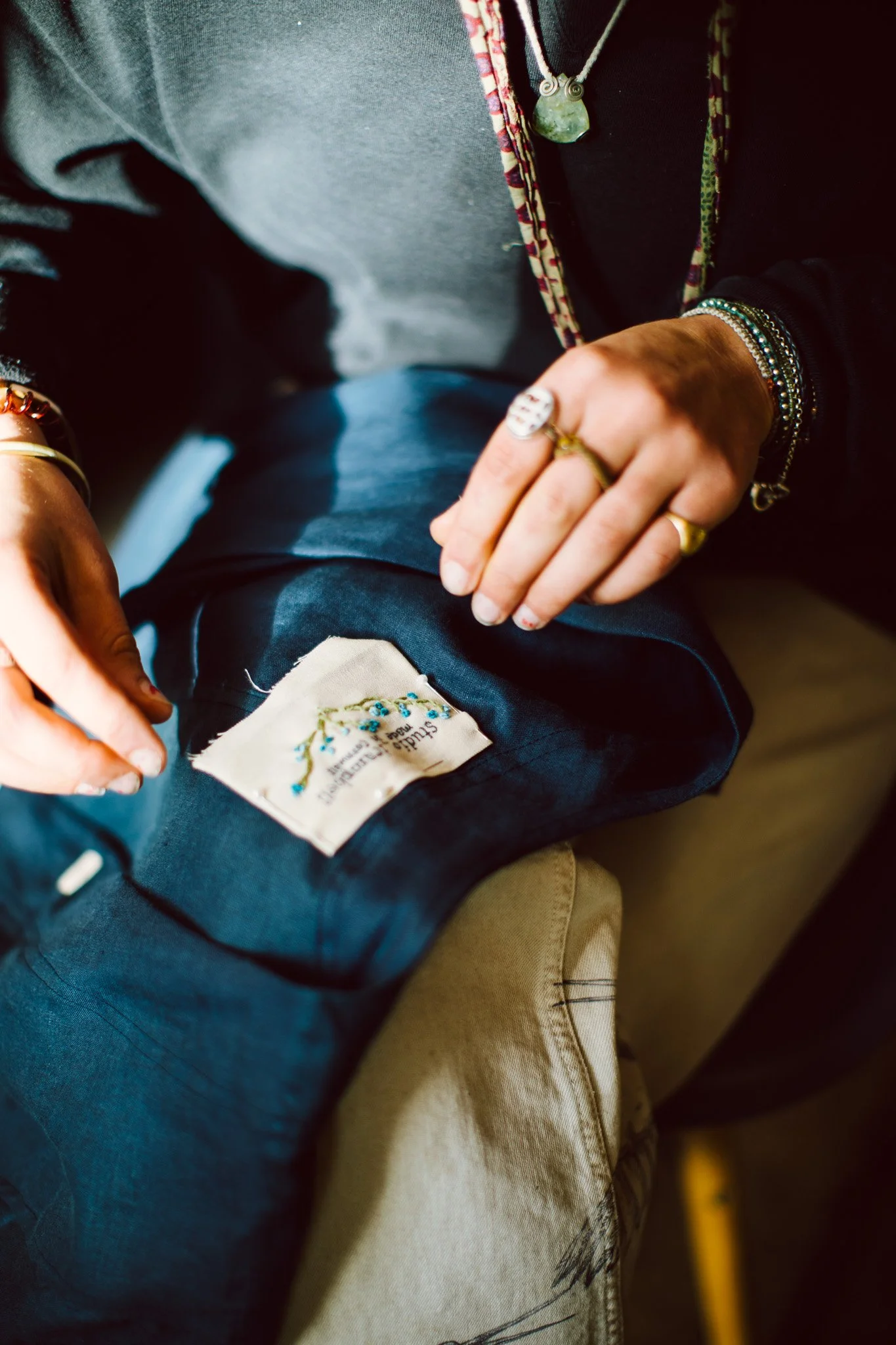 A person wearing jewelry and a gray top sitting on a beige couch with a dark blue blanket with a label on it, holding some embroidered fabric or jewelry in their hand.