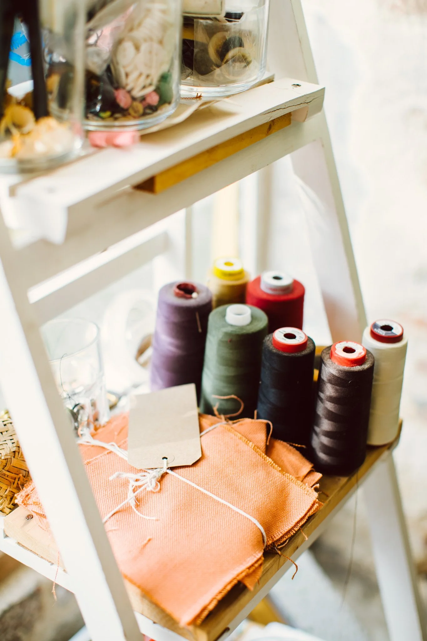 Spools of sewing thread in various colors, orange fabric, and a glass of water on a small wooden and white shelf.