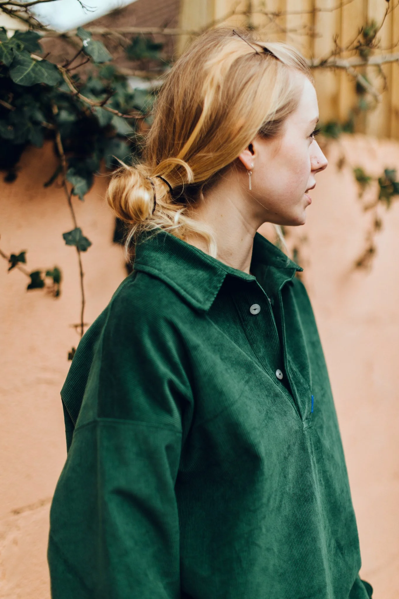 A woman with blonde hair in a loose bun, wearing a dark green corduroy shirt, standing sideways against a background of a beige wall with green ivy.
