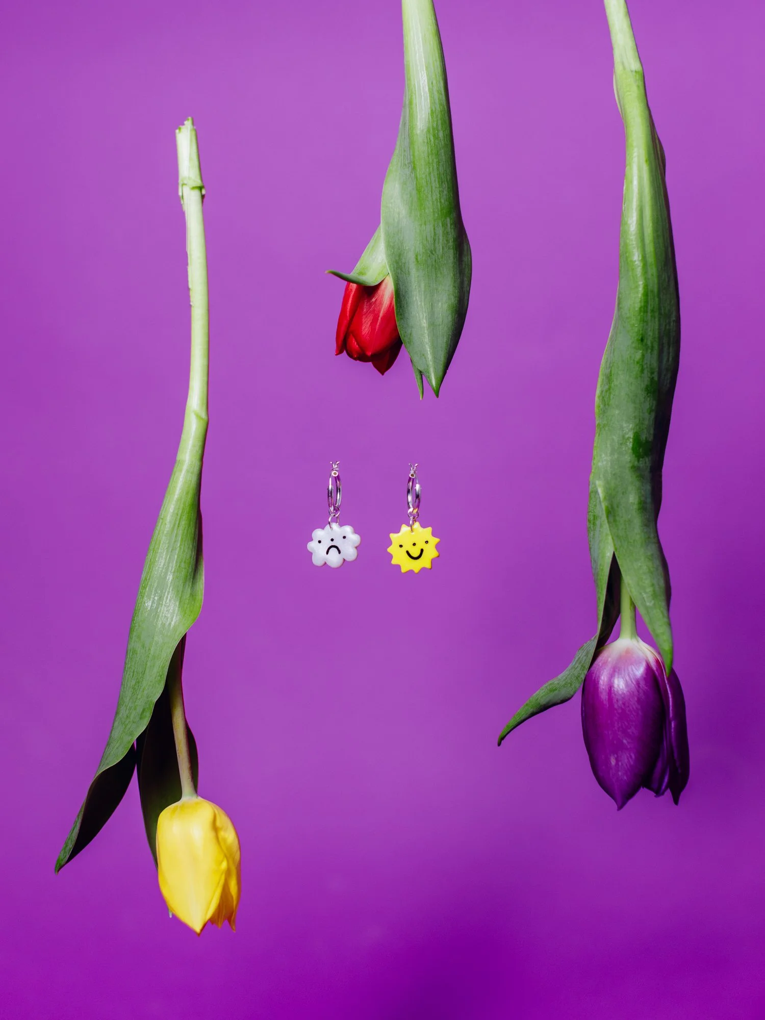 Three tulips in yellow, purple, and red with green stems and leaves hanging upside down against a purple background. Two earrings hanging between the tulips, one with a white sad cloud and the other with a yellow smiling sun.