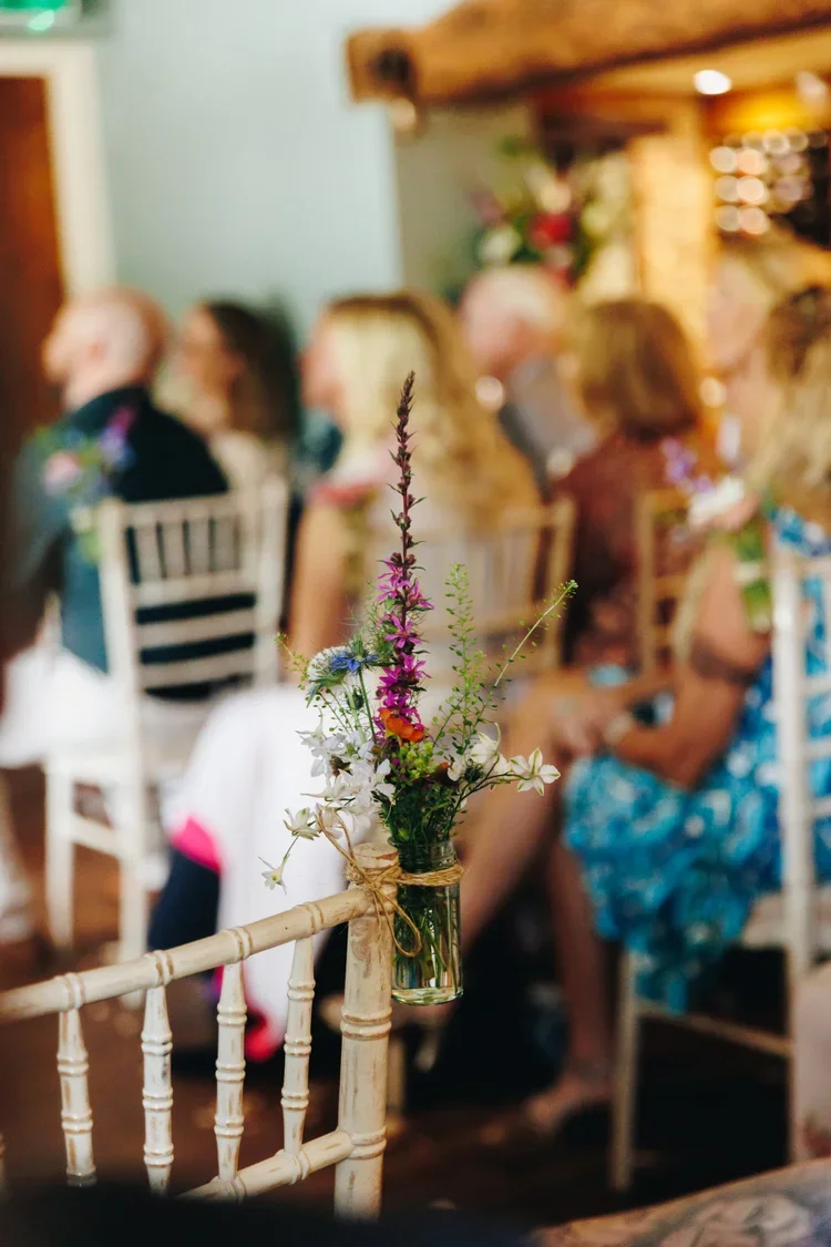 A small glass jar with wildflowers hanging on a white wooden chair at a gathering, with blurred people sitting in the background.