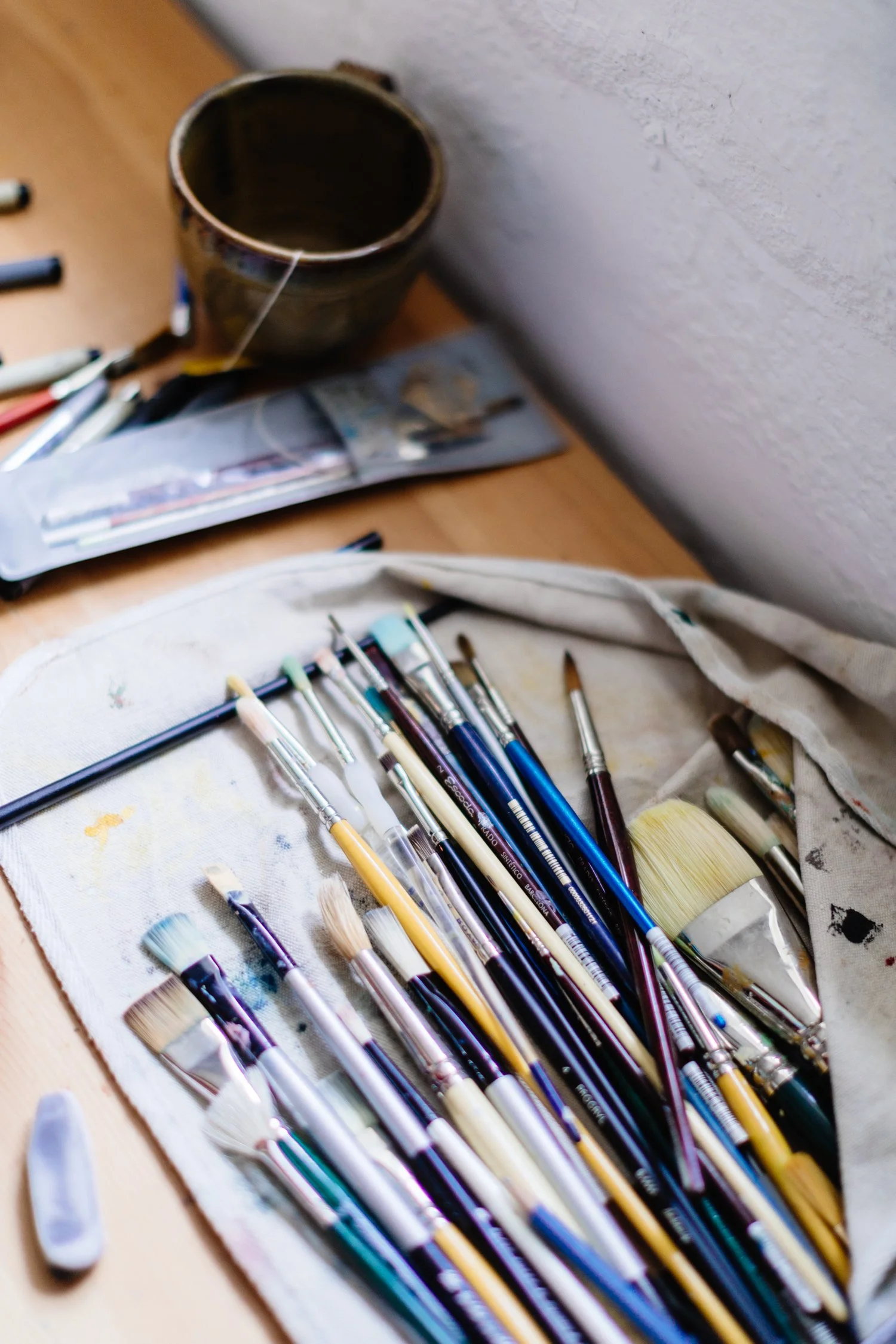 A collection of paintbrushes in a fabric holder, with an empty cup, pens, and a plastic pouch on a wooden table.